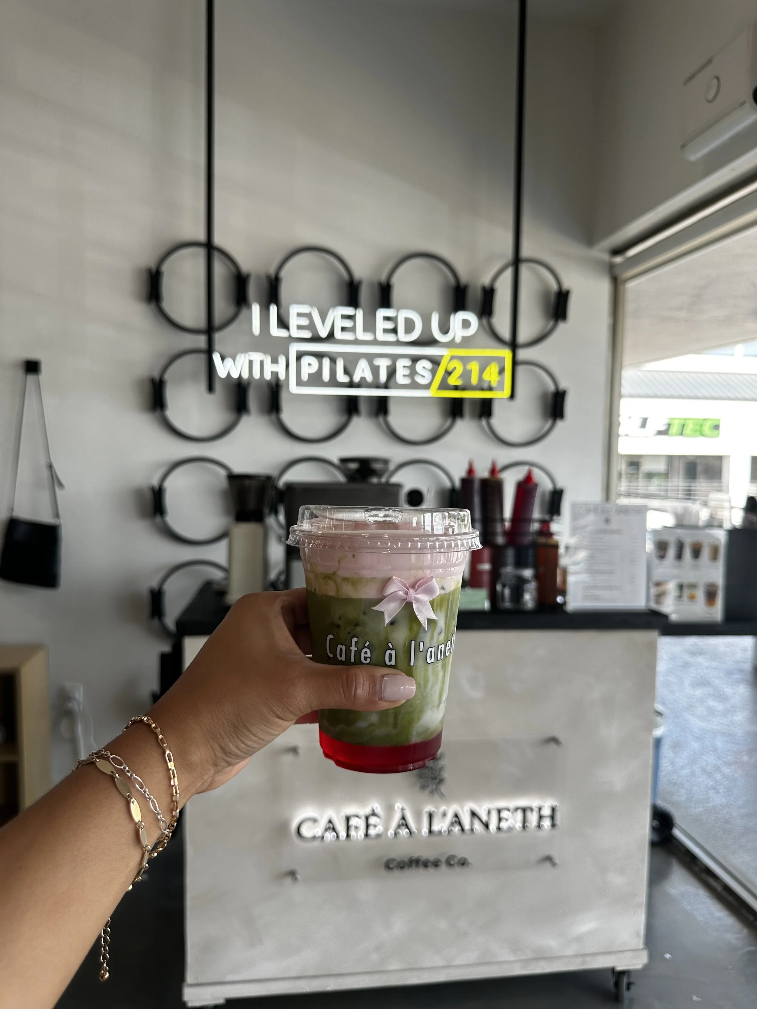 A hand holding a layered green and pink drink with a pink bow on a clear cup at Café à l'Aneth, with a sign that says 'I leveled up with Pilates 214' in the background.
