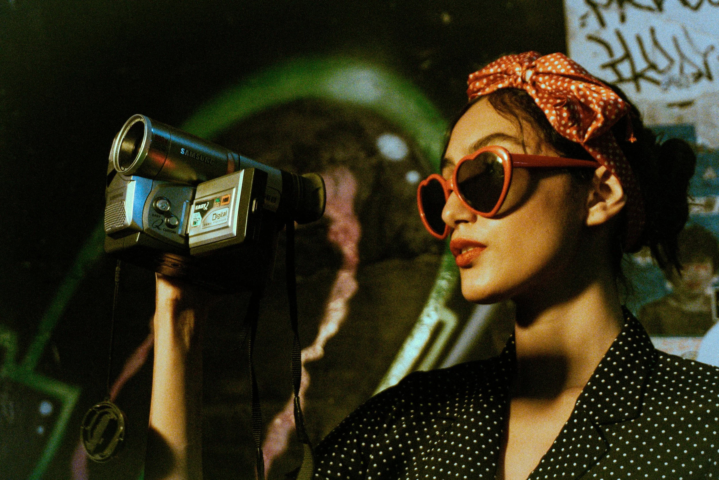 A woman wearing heart-shaped sunglasses and a polka dot shirt, with a headscarf, is looking through a vintage camera mounted on a stand, with graffiti and colorful art in the background.