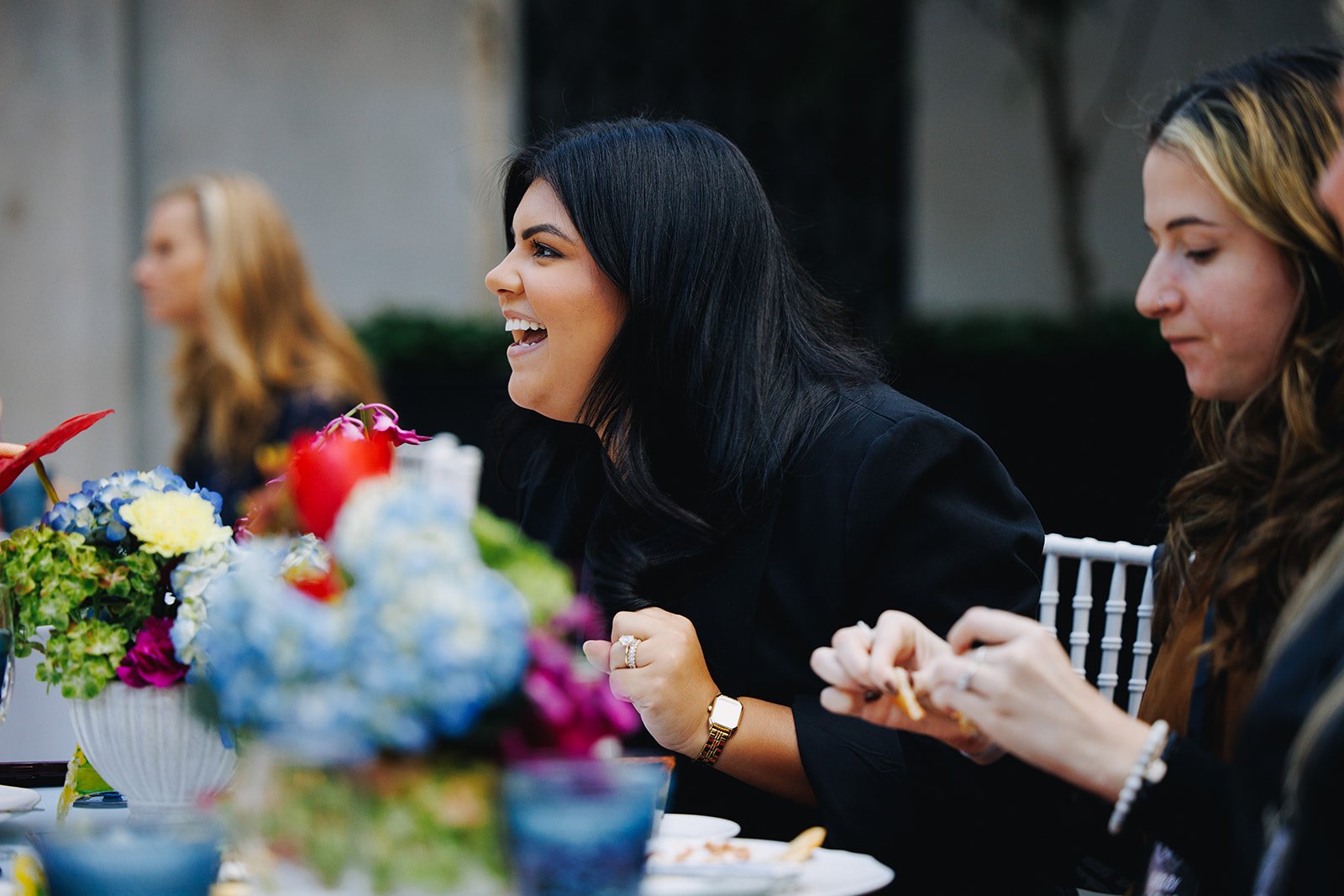 A woman with black hair smiling and talking at a meal with others. There are colorful flower arrangements on the table.