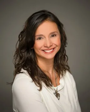 A woman with curly dark hair, smiling, wearing a white top and a necklace, against a gray background.