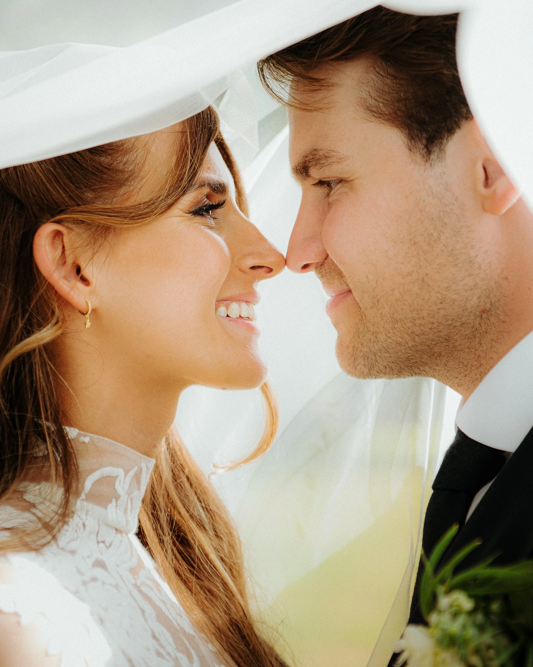 Close-up of a bride and groom smiling with their noses almost touching, under a wedding veil.