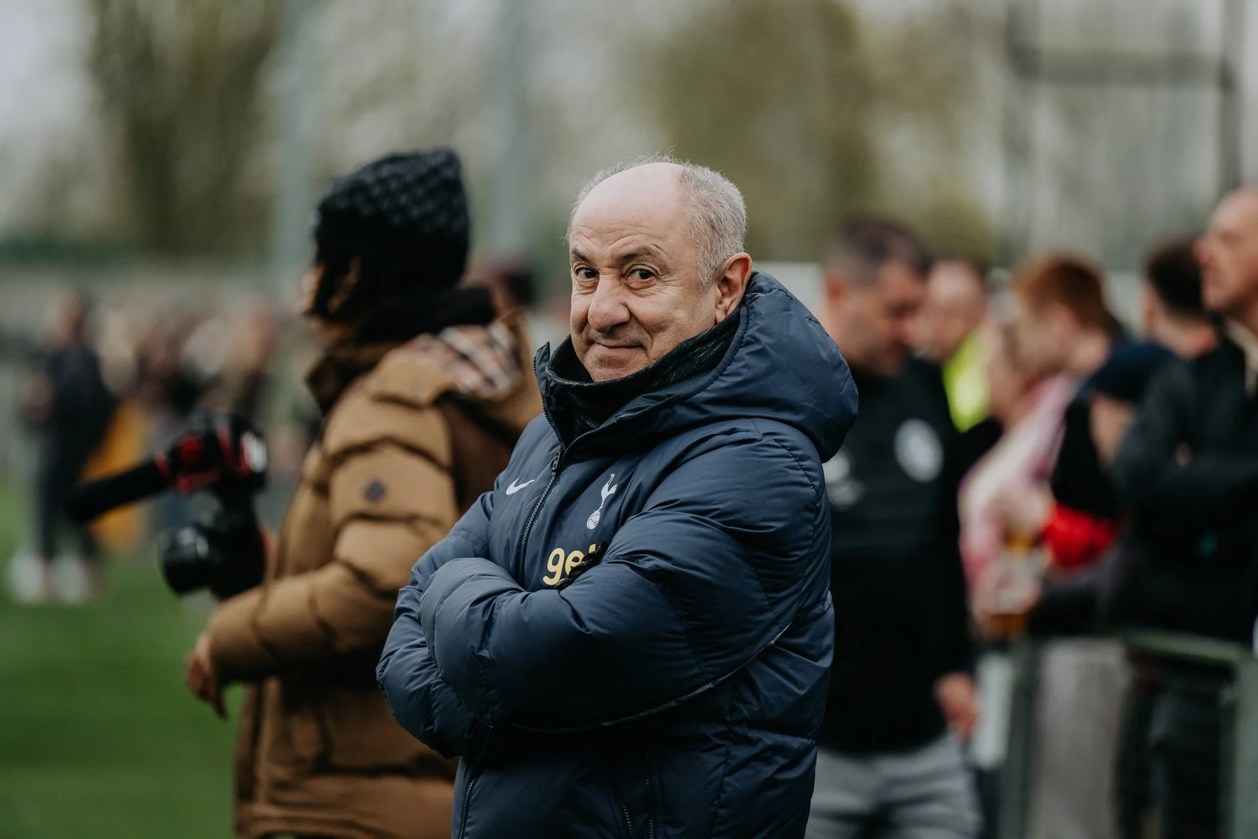 A man in a navy blue jacket with the Tottenham Hotspur logo standing with his arms crossed, looking at the camera with a slight smile, surrounded by a crowd of people at an outdoor event