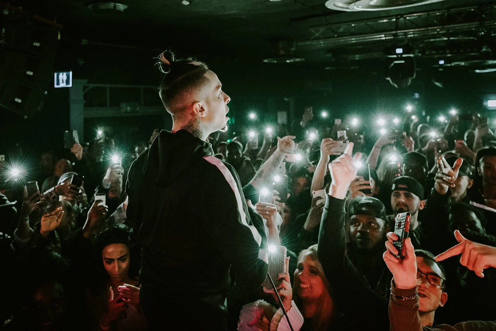 A performer with a man bun hairstyle sings in front of a crowd taking photos and videos with their smartphones at a concert.