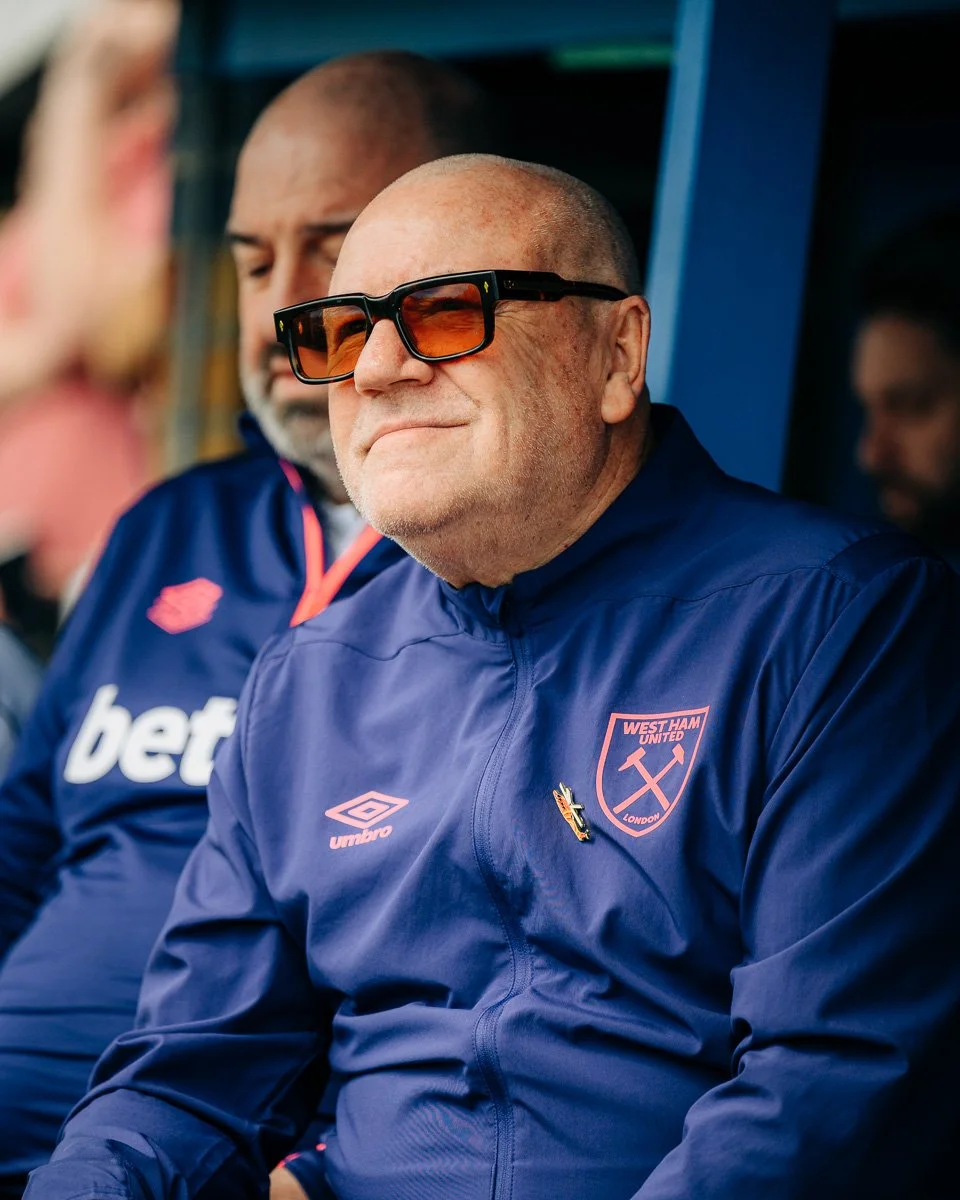 A man wearing dark sunglasses and a West Ham United football jacket sitting on a team bench.