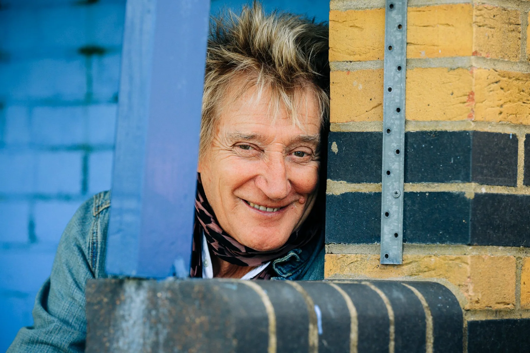 A smiling woman with blonde hair peeking through a gap between a blue door and a brick wall.