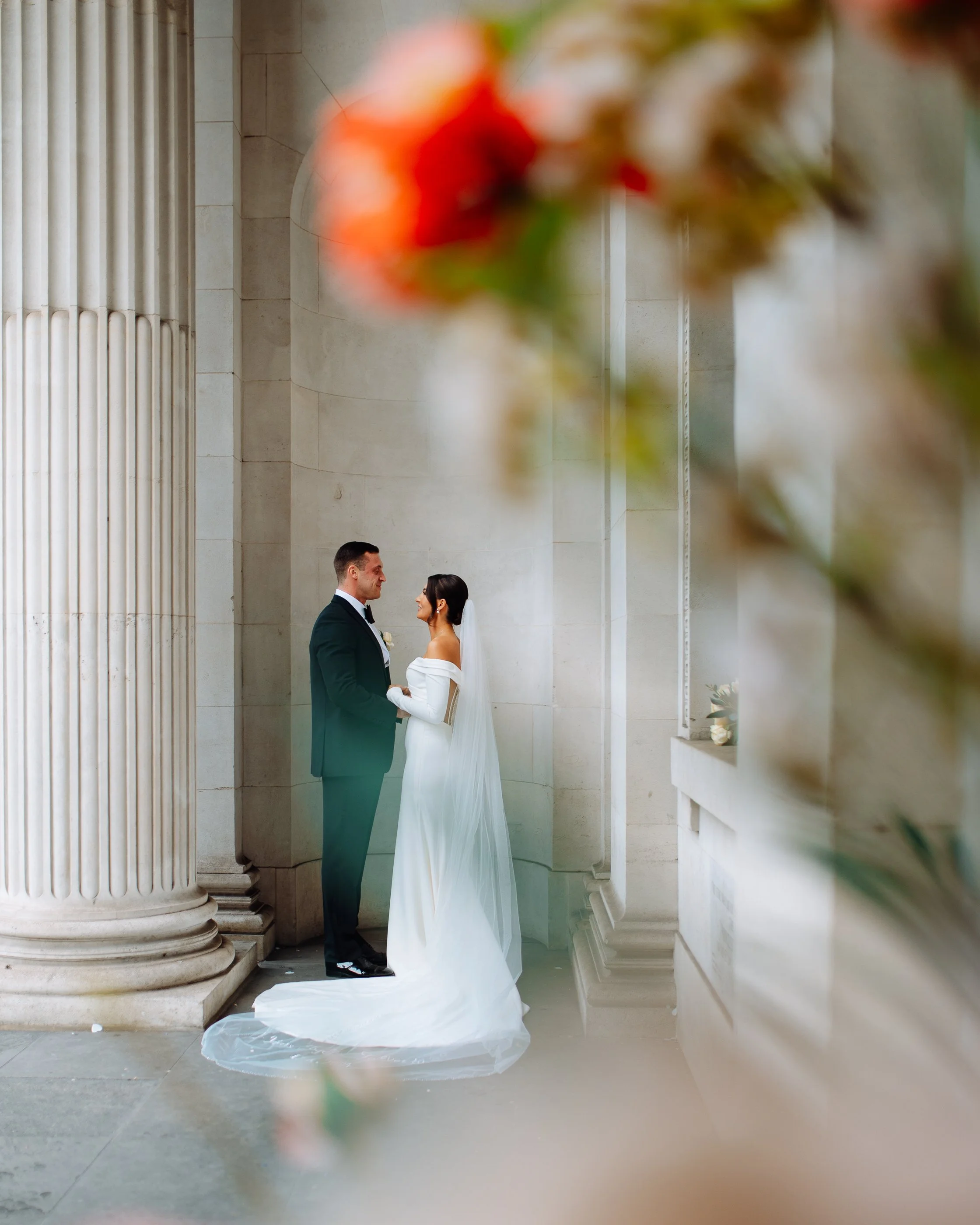 Bride and groom standing close together in front of a classical stone building with tall columns, holding hands and looking at each other, with out-of-focus flowers in the foreground.