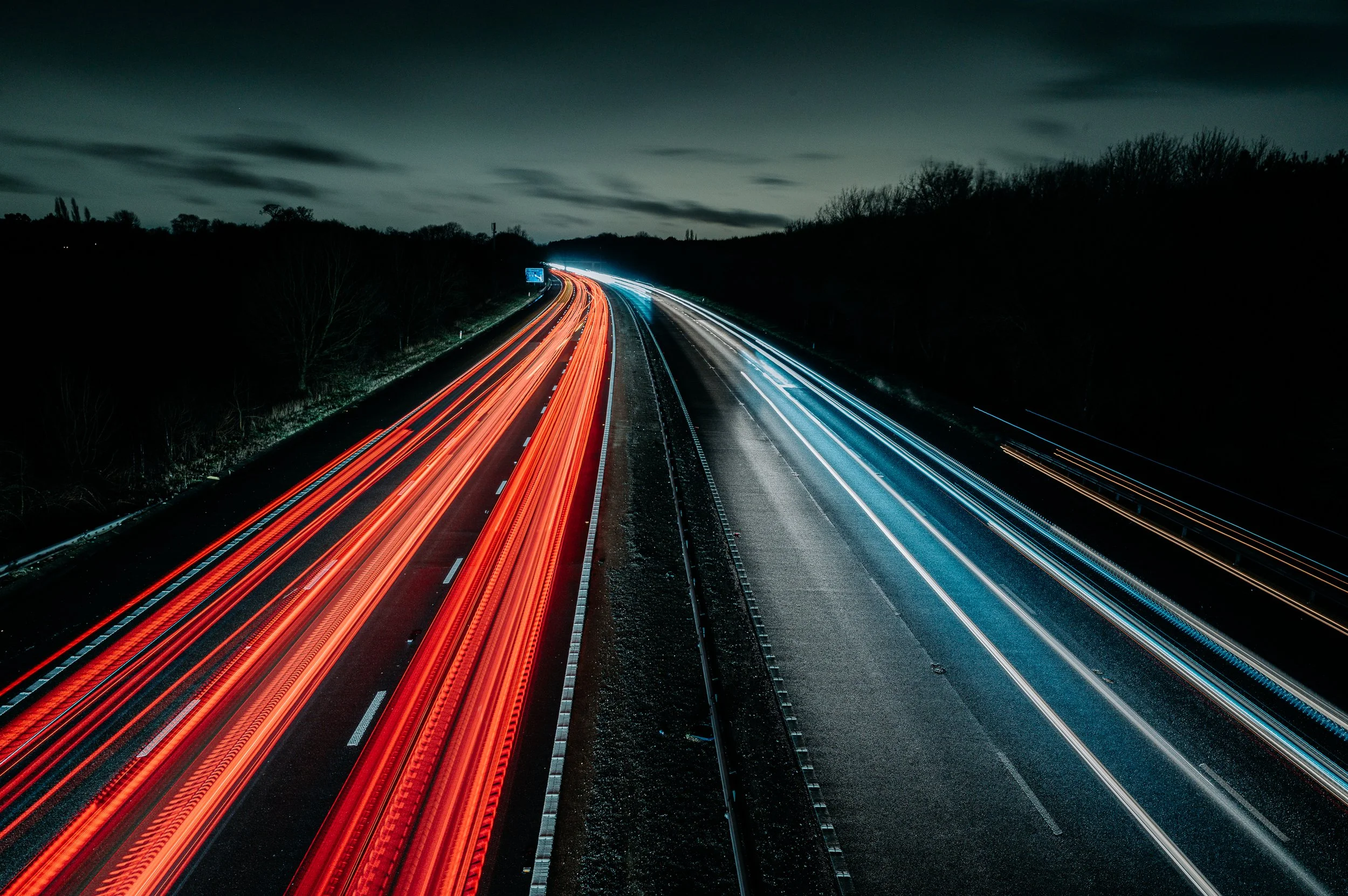 Long exposure photo of a highway at night showing red and white light trails from vehicles, with dark trees and cloudy sky in the background.
