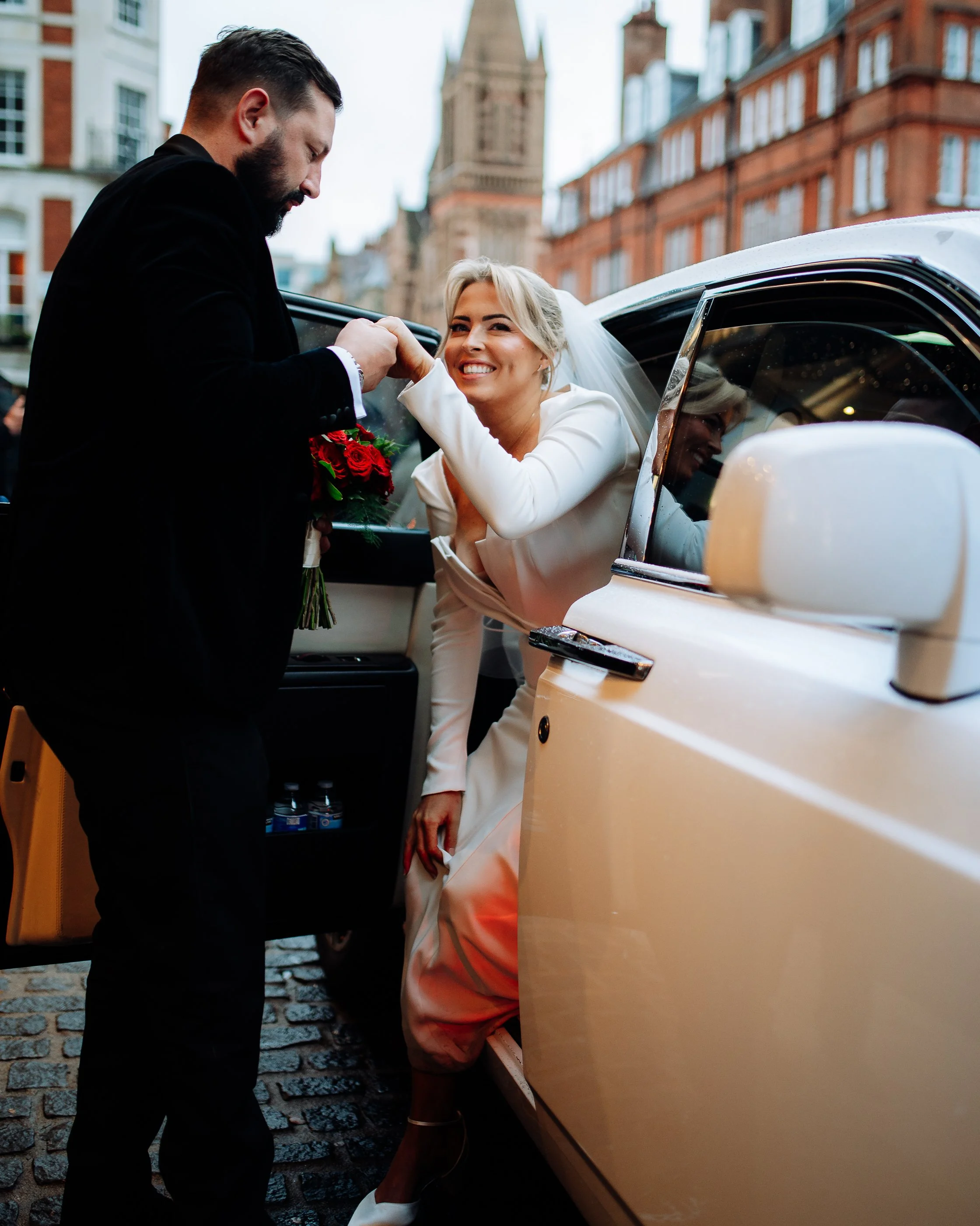A bride in a white dress smiling as she interacts with a man in a black suit next to a white vintage car, with city buildings in the background.
