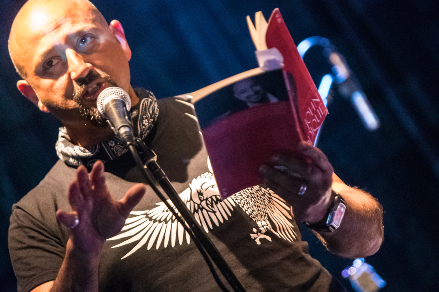 Author John Siddique wearing a black t-shirt with an eagle design, reading from his book Full Blood during a live performance.