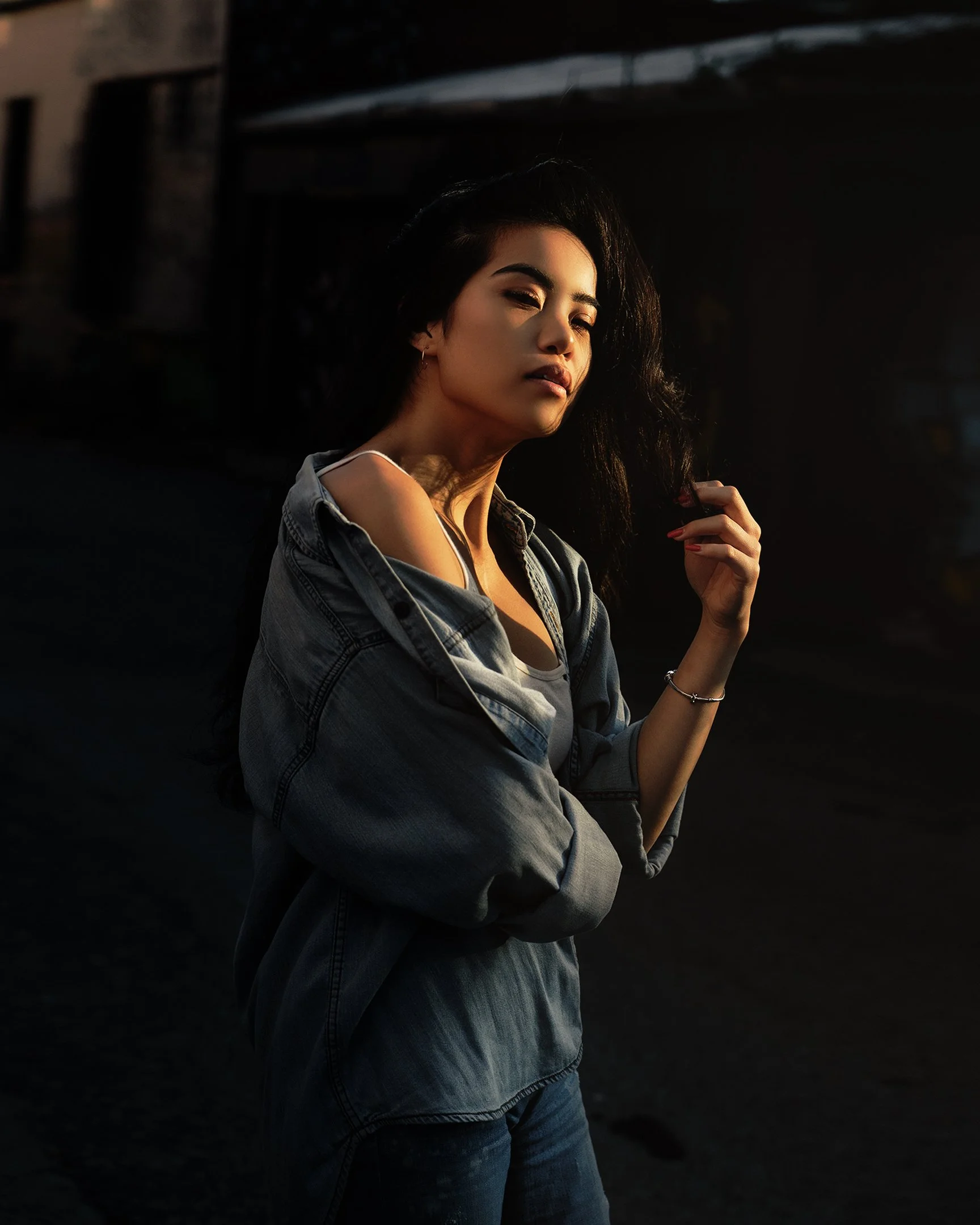 A young woman with dark hair and tan skin poses in sunlight wearing a denim jacket and white top, with a dark background.
