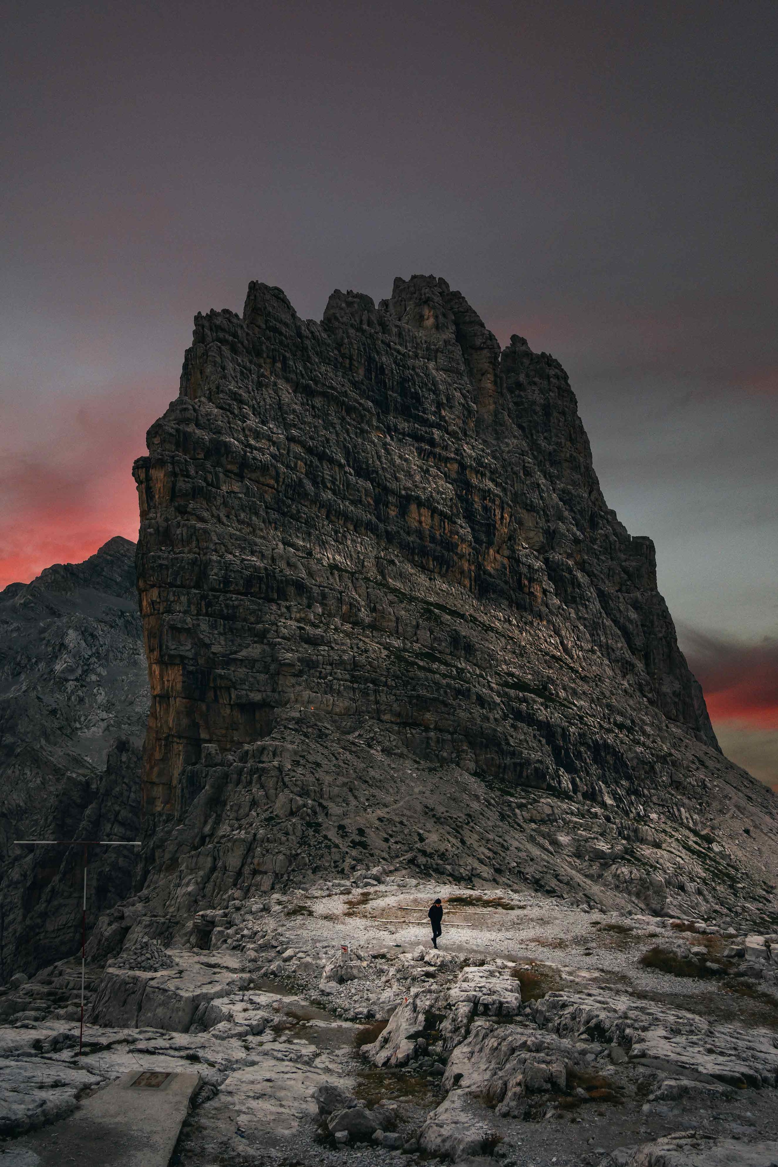 A lone person standing at the base of a tall, rugged mountain at dusk, with a colorful sky in the background.