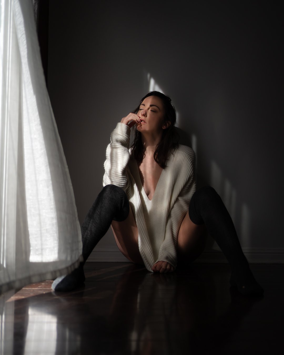 A woman sitting on the floor near a window with white curtains, wearing a loose beige sweater and black knee-high socks, with sunlight casting a dramatic shadow on the dark wall behind her.