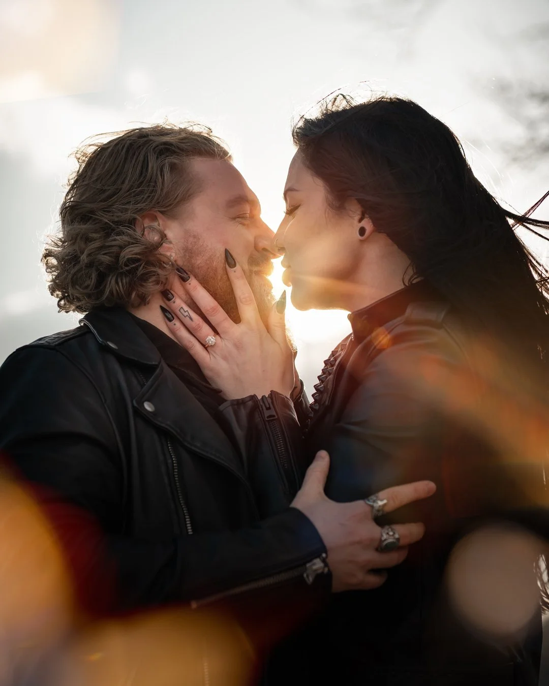 A couple is close together, smiling and about to kiss, with the sun shining behind them and a blurred background.
