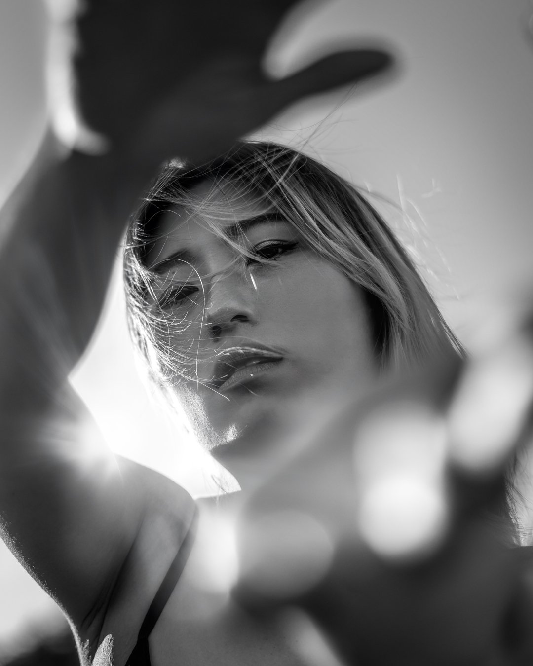Close-up black and white photo of a woman with short hair, partially obscured by her arm and hand, looking downward with sunlight highlighting her face and hair.