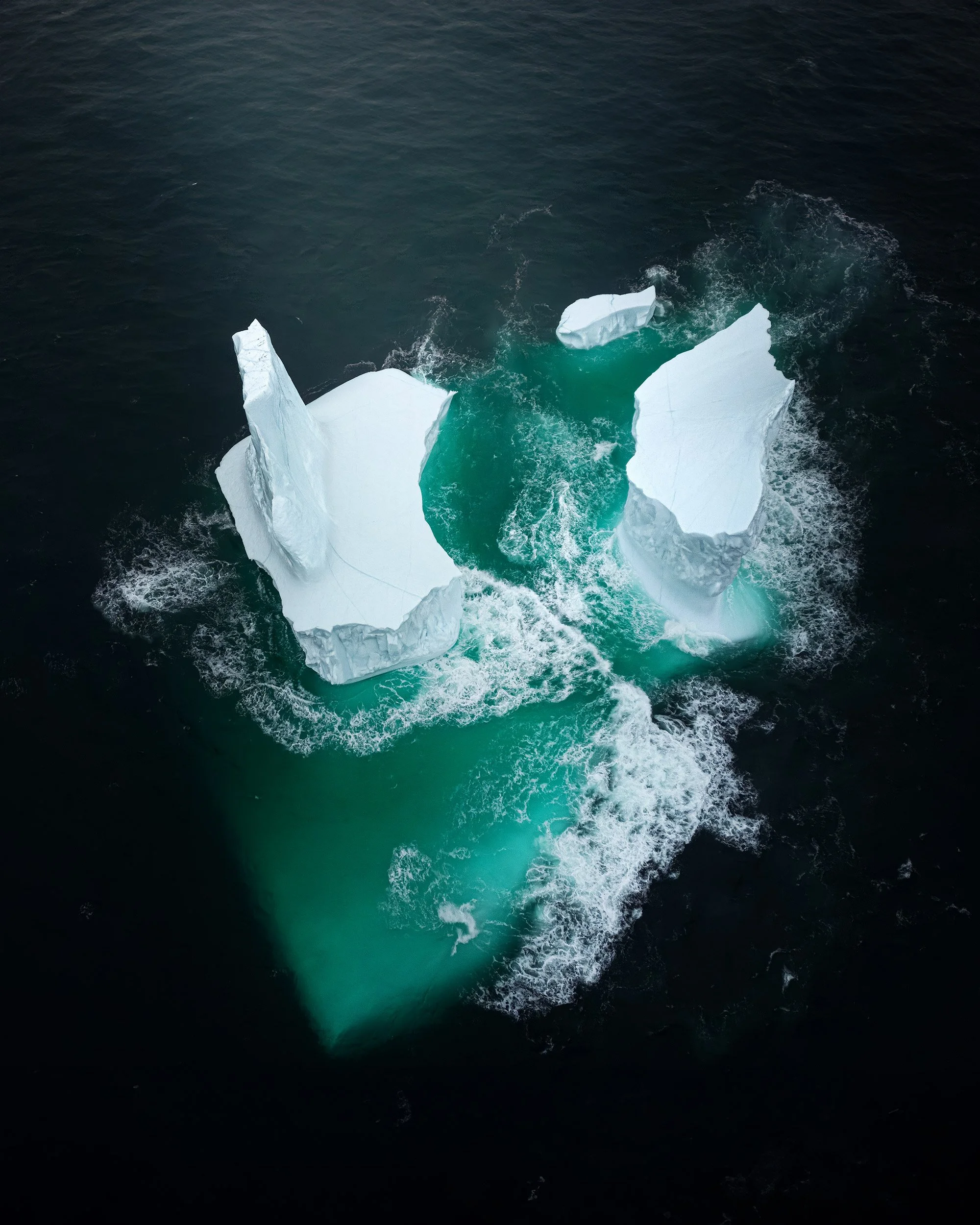 Aerial view of a large iceberg floating in the ocean, with visible cracks and a deep turquoise center, surrounded by dark water.