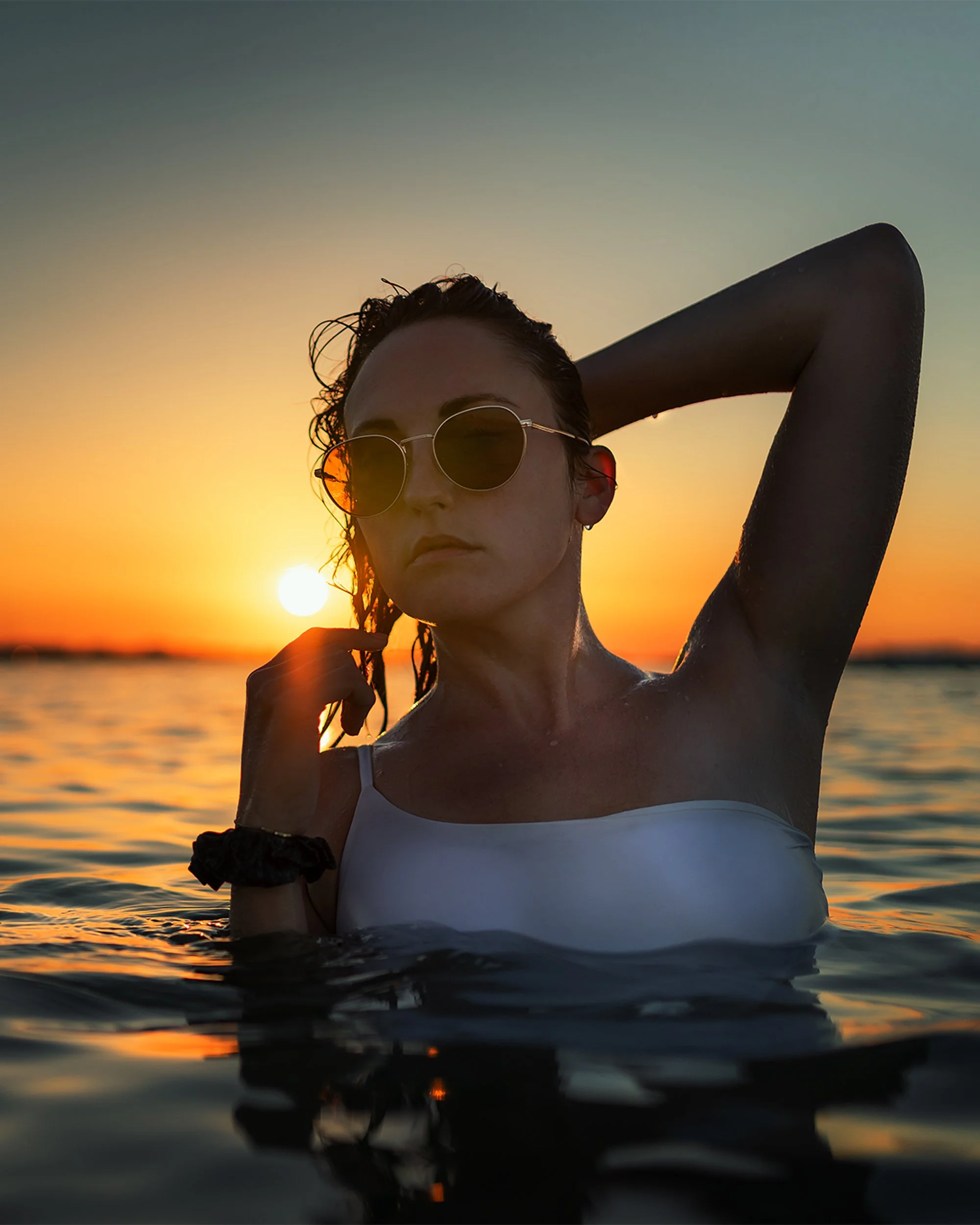 A woman wearing sunglasses and a white swimsuit, standing in water during sunset with her arm raised behind her head.