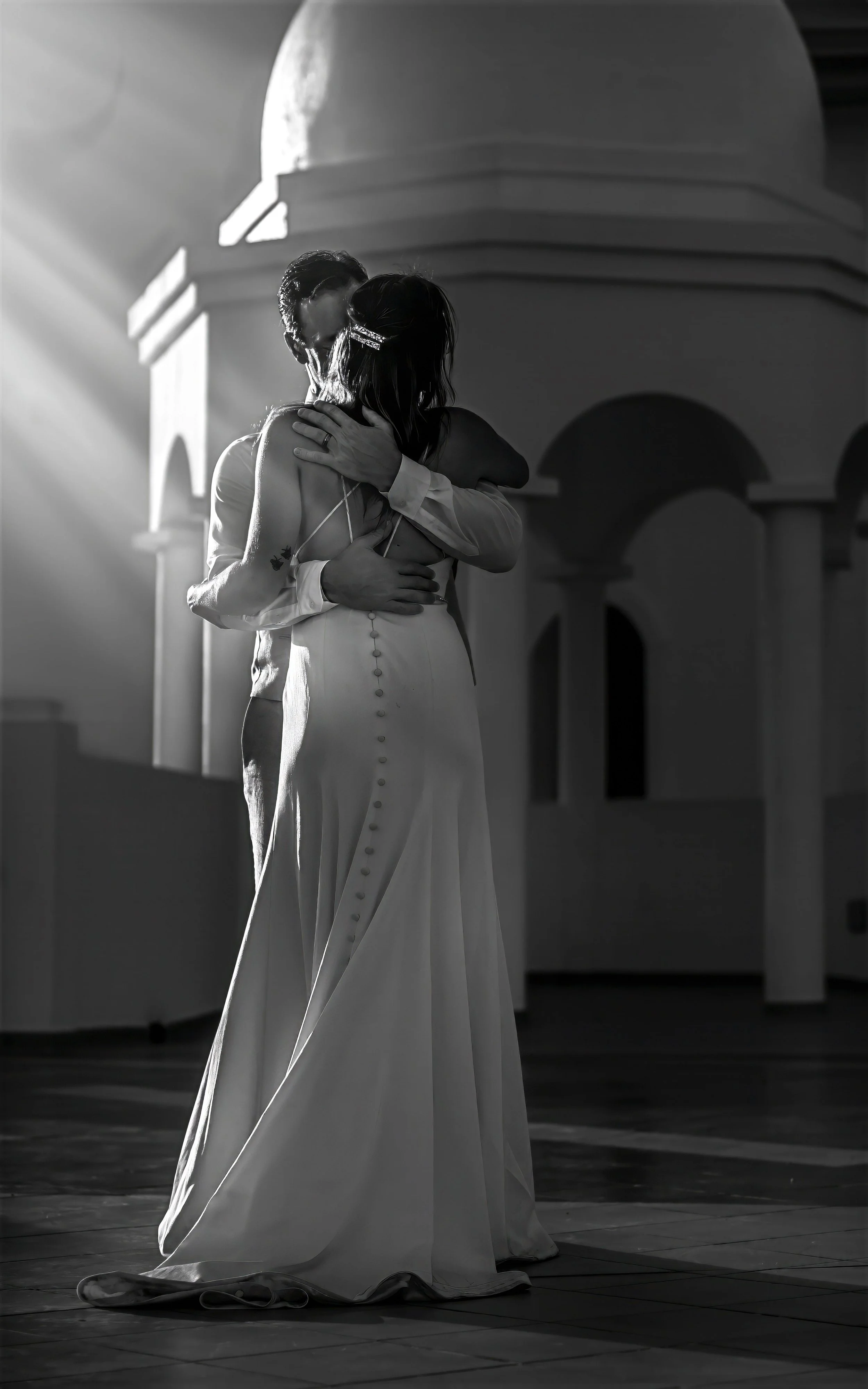 A couple dancing closely on a dance floor, backlit by a bright light, with architecture featuring arches and domes in the background.