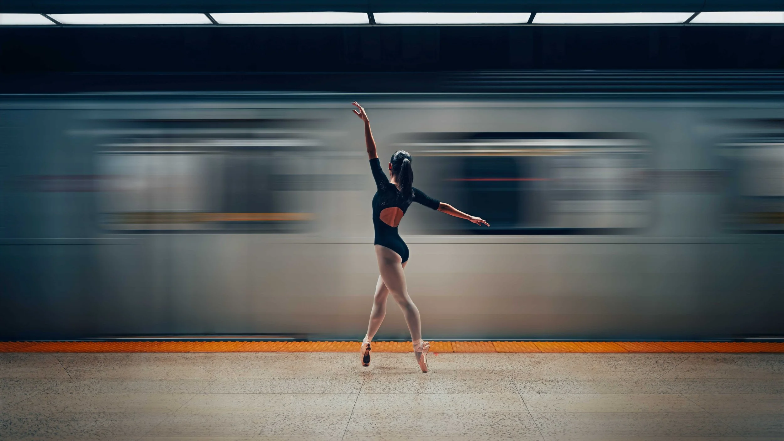 Ballet dancer in black leotard and pink pointe shoes performing ballet pose on subway platform with a moving train in background.