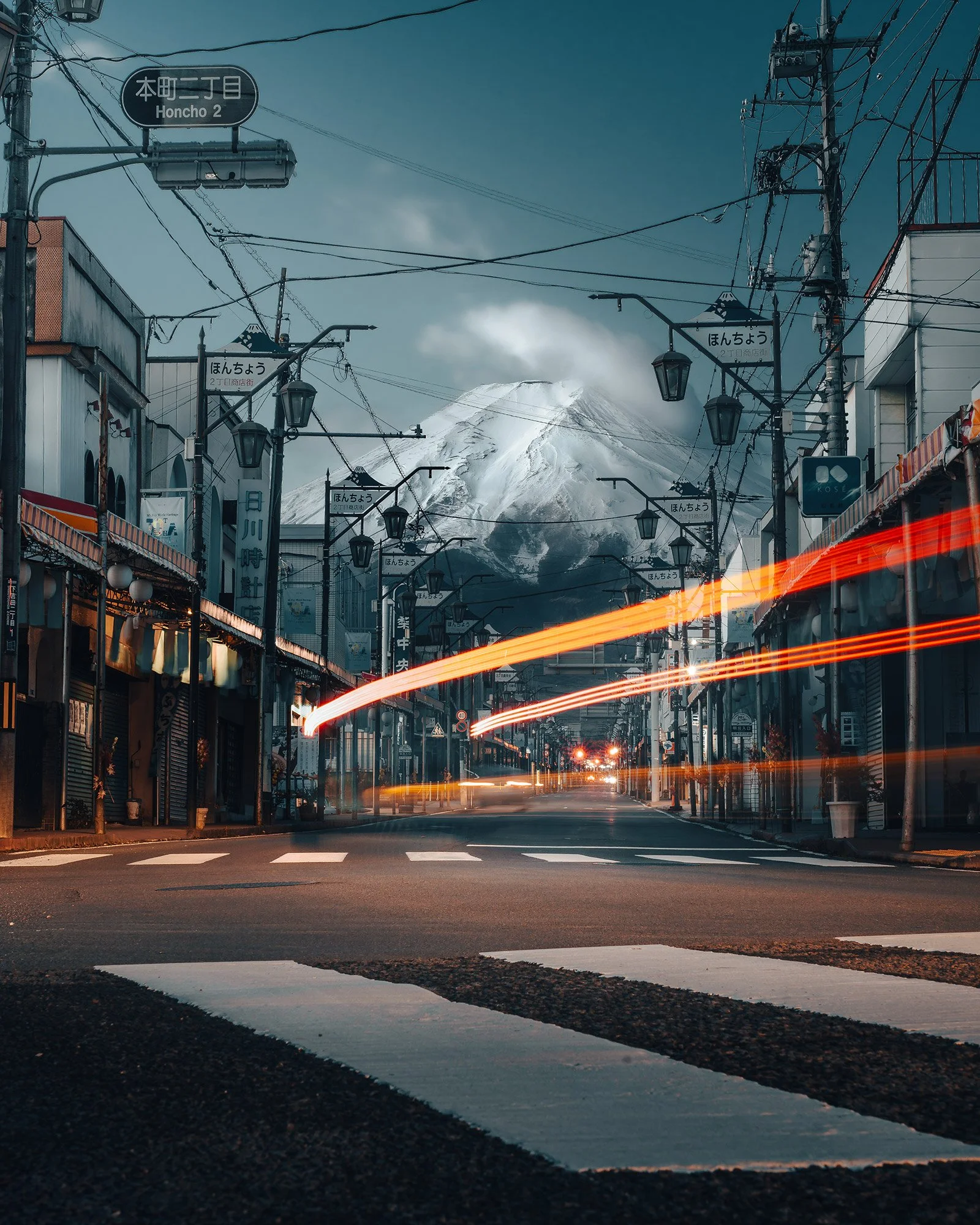 Empty street with shops and streetlights leading towards a snow-capped mountain, possibly Mount Fuji, in the background, during dusk with light streaks from passing vehicles.