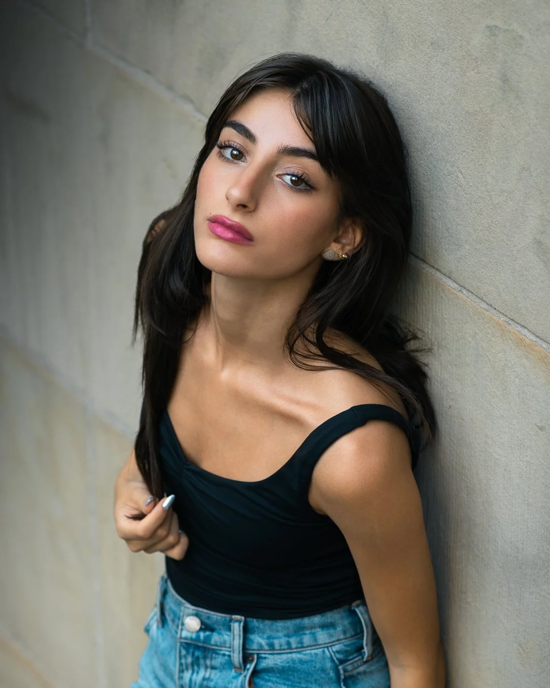 A young woman with long dark hair, wearing a black sleeveless top and blue jeans, leaning against a beige wall, looking at the camera.