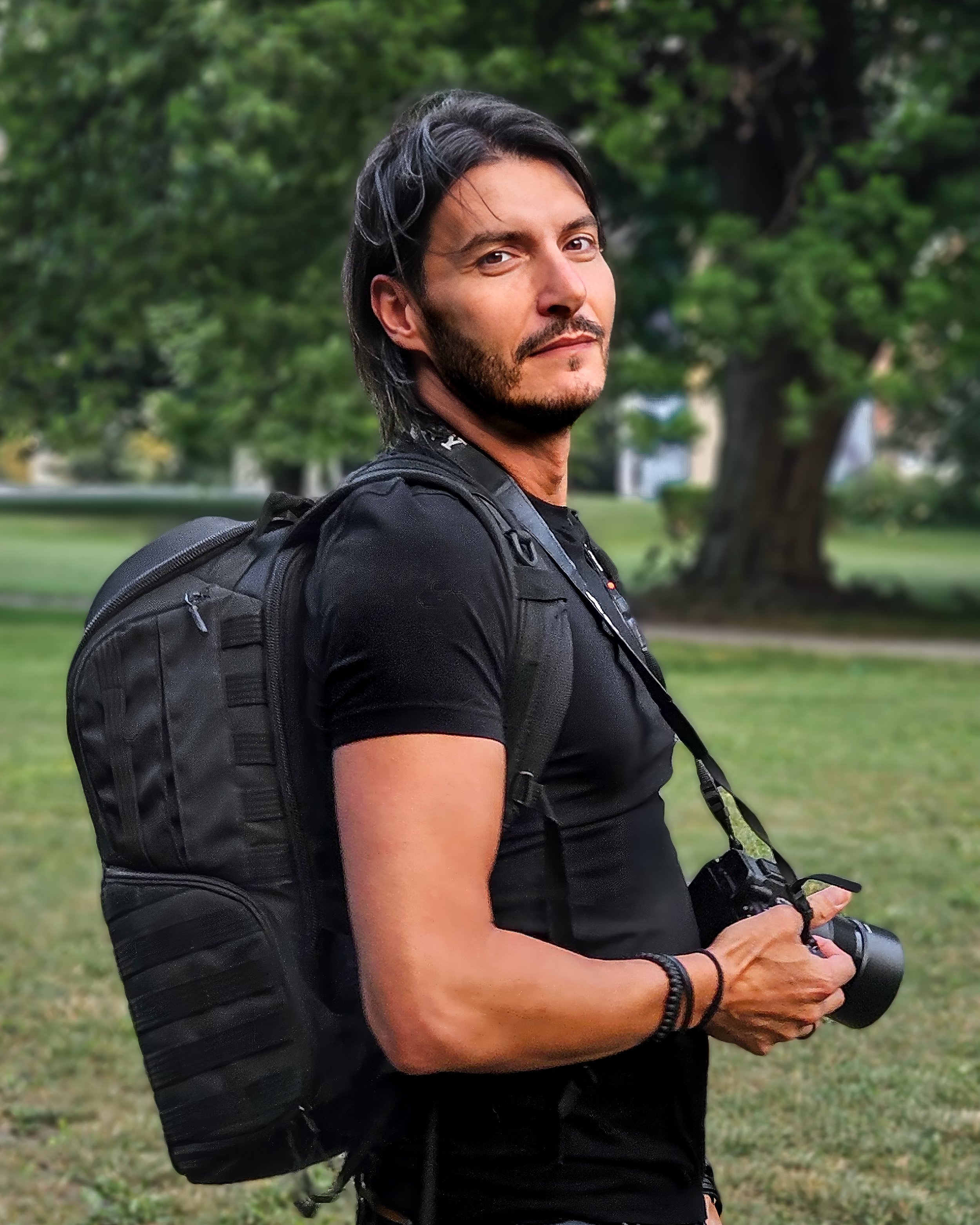 Man with dark hair and beard standing outdoors, carrying a black backpack and holding a camera, with trees in the background.