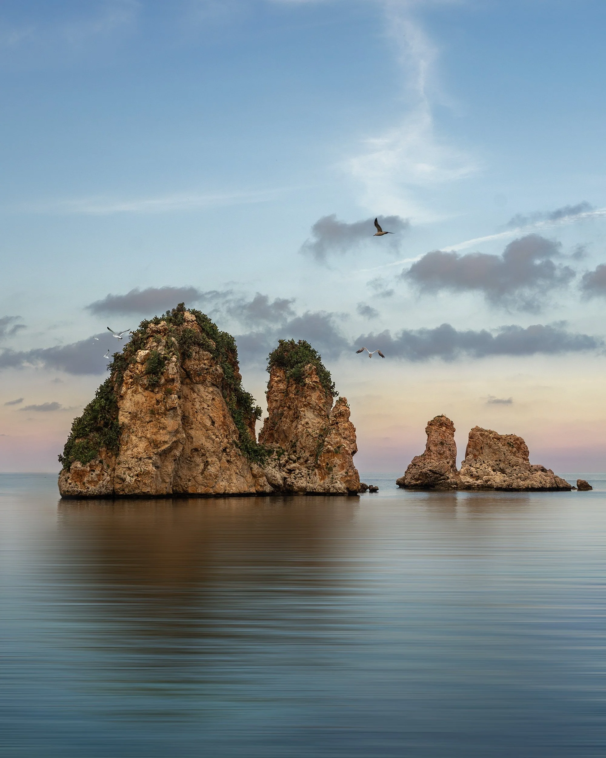 Seaside scene with three large rocky islands in calm water, a sky with clouds, and several seagulls flying overhead during sunset or sunrise.