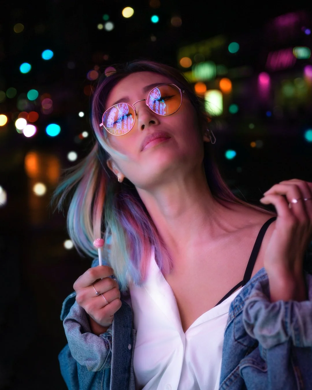 A young woman with rainbow-colored hair wearing sunglasses reflecting neon lights, holding a lollipop, in an urban night setting with colorful bokeh lights.