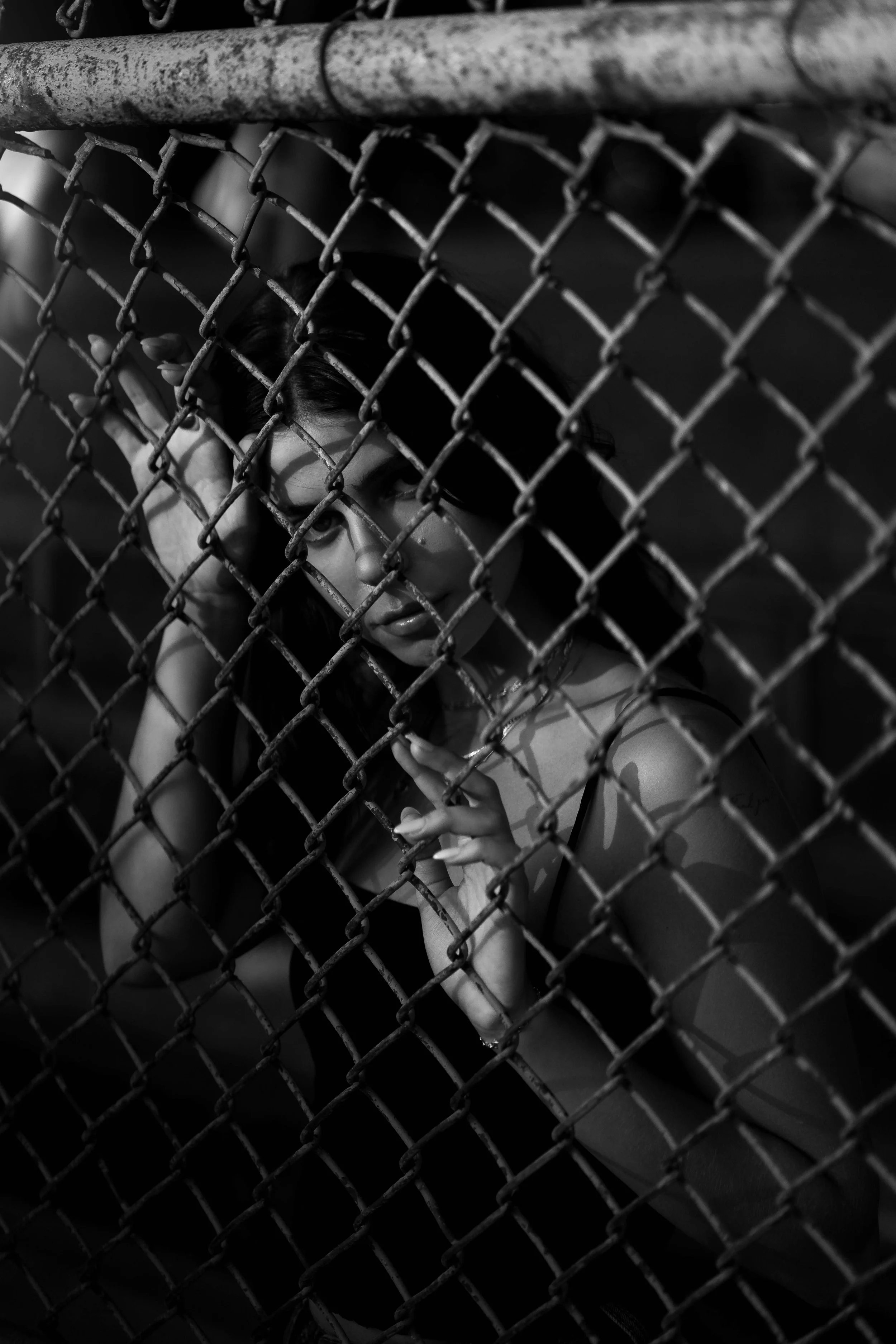 A woman posing behind a chain-link fence, with her hand and face pressed against the fence, in a black and white photograph.