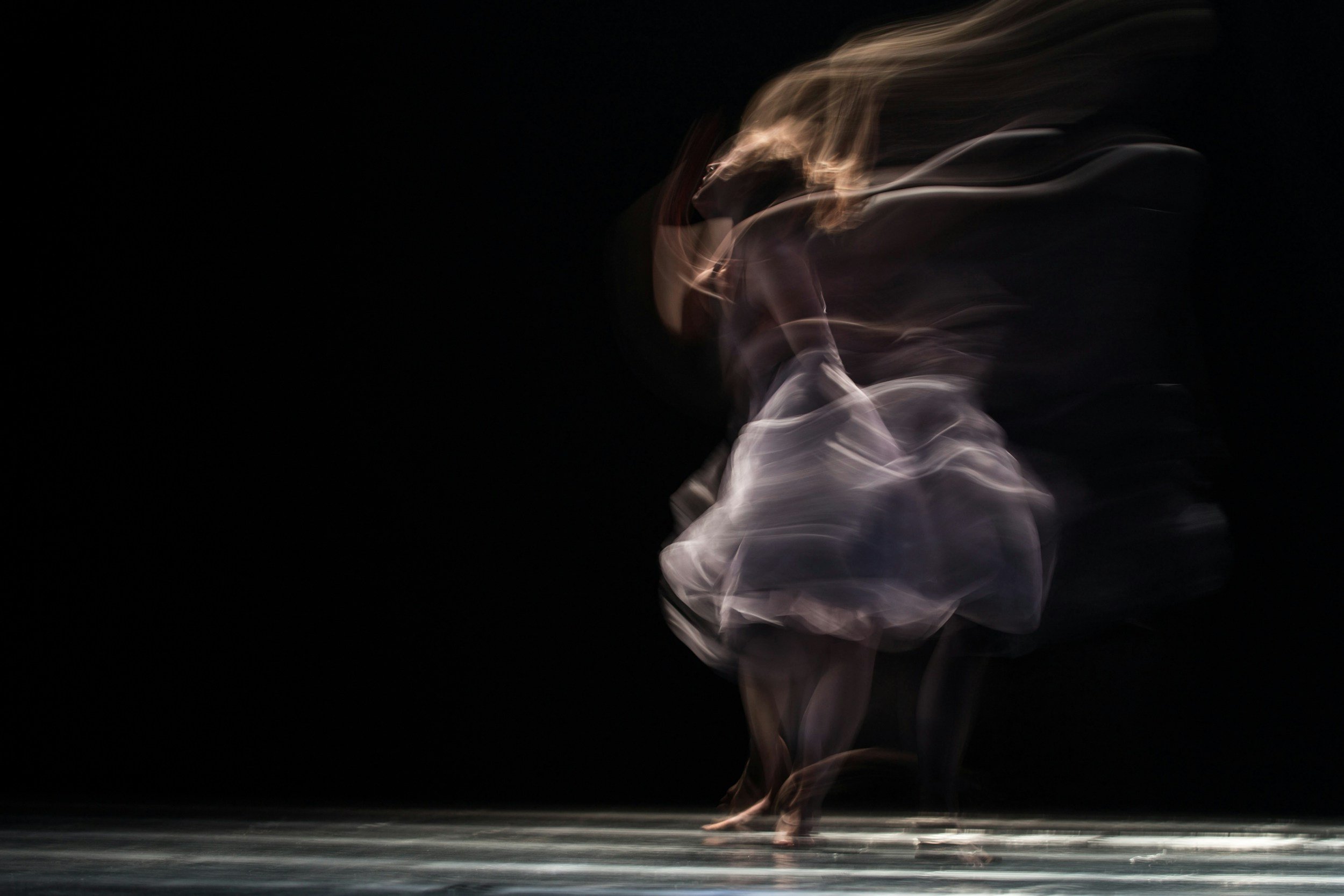 A dancer in motion with a flowing dress on a dark stage, creating a blurred, ghostly effect.