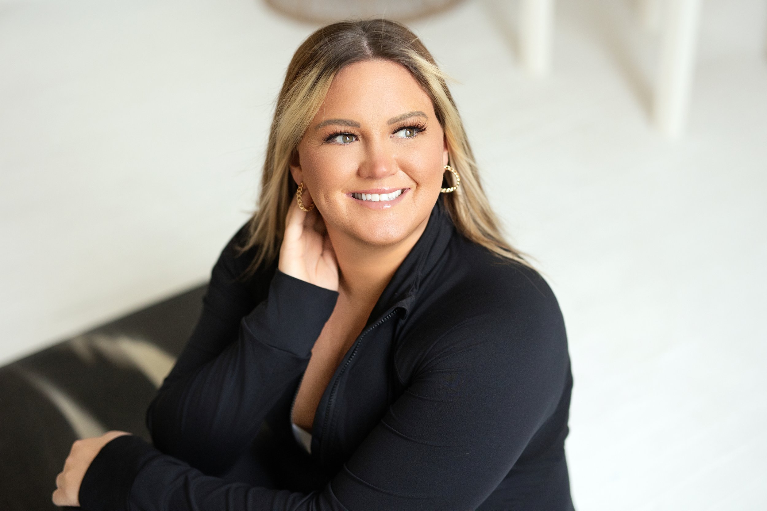 A smiling woman with blonde hair, wearing a black jacket and gold earrings, sitting on a black leather couch against a white wall.