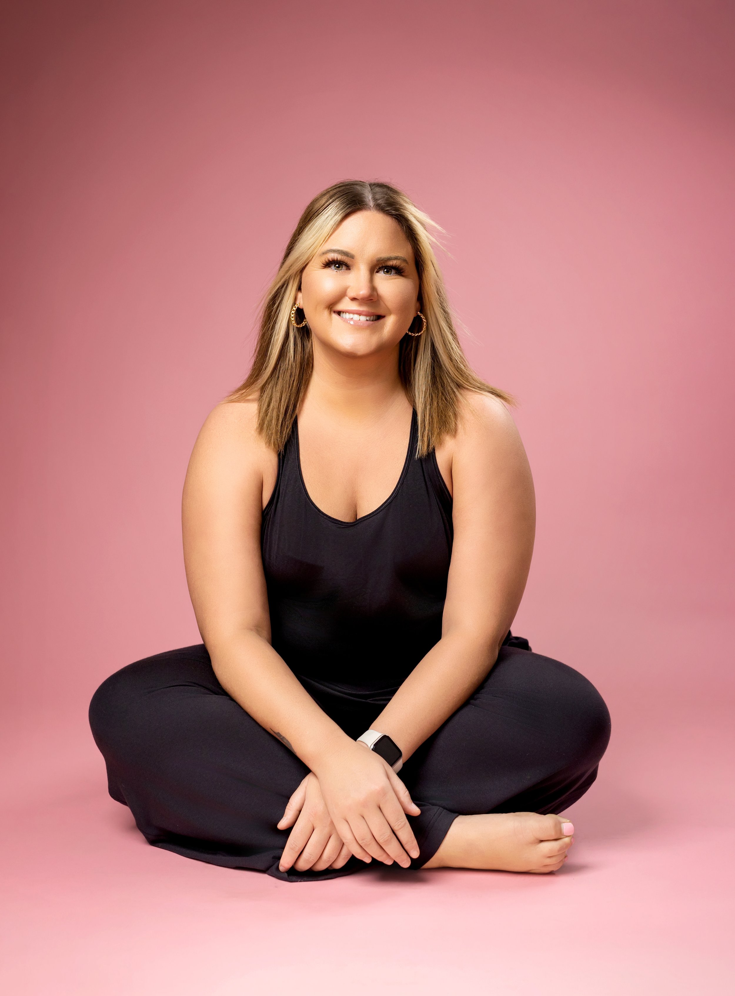 A woman sitting cross-legged on a pink background, smiling, wearing a black sleeveless top and black pants, with earrings and a smartwatch.
