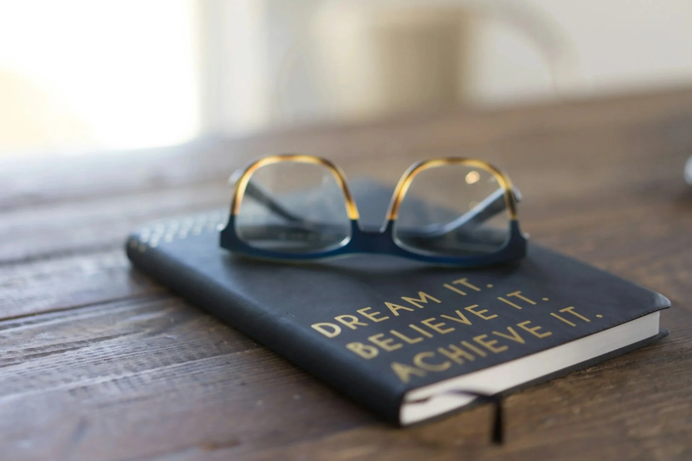 A pair of eyeglasses resting on a black notebook with gold text that says "DREAM IT. BELIEVE IT. ACHIEVE IT." on a wooden surface.