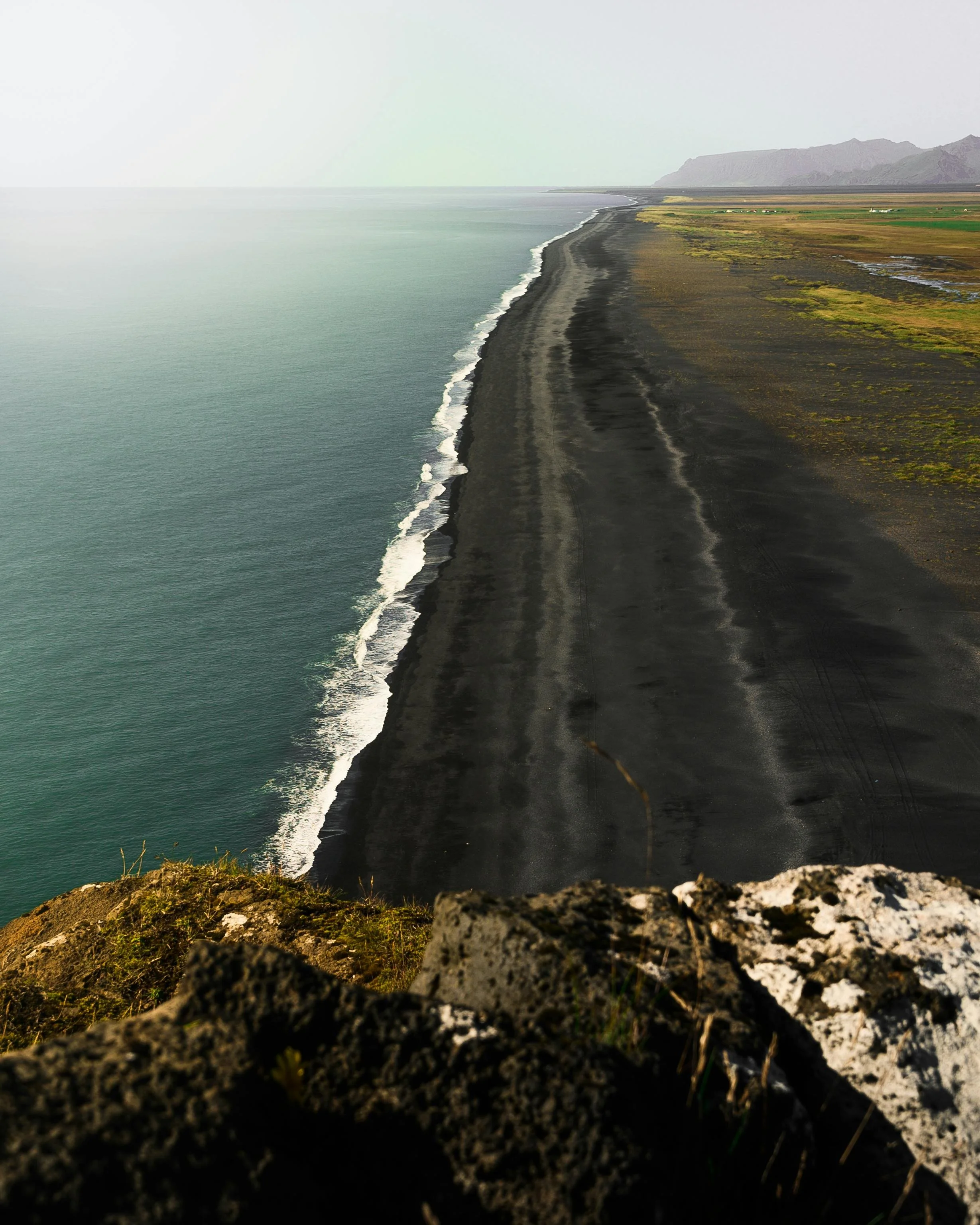 Aerial view of a black sand beach along the coastline, with green fields and mountains in the background.
