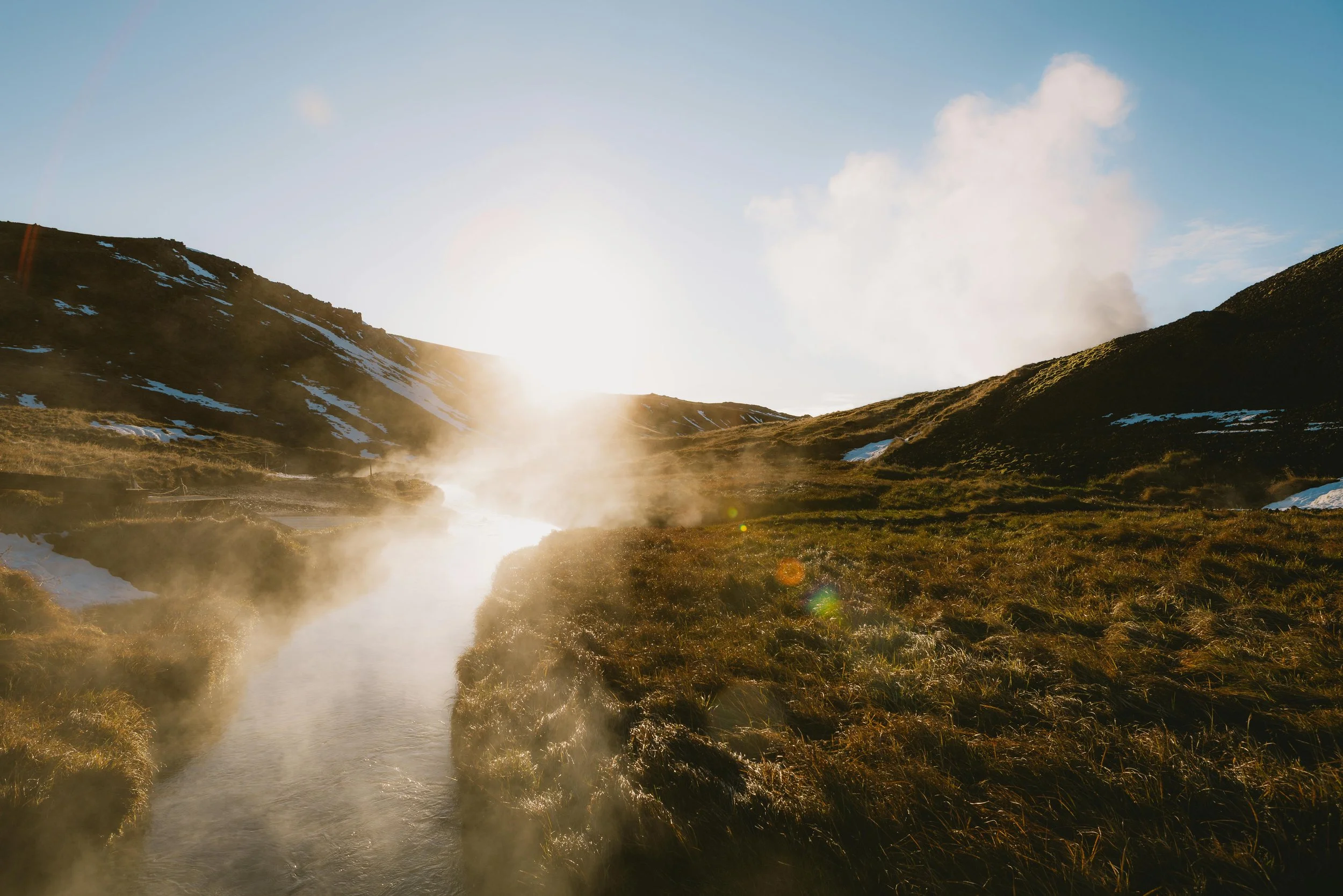 A scenic landscape of a mountain valley with a river flowing through it, surrounded by grassy hills, with the sun rising or setting in the background and steam or mist rising from the water.