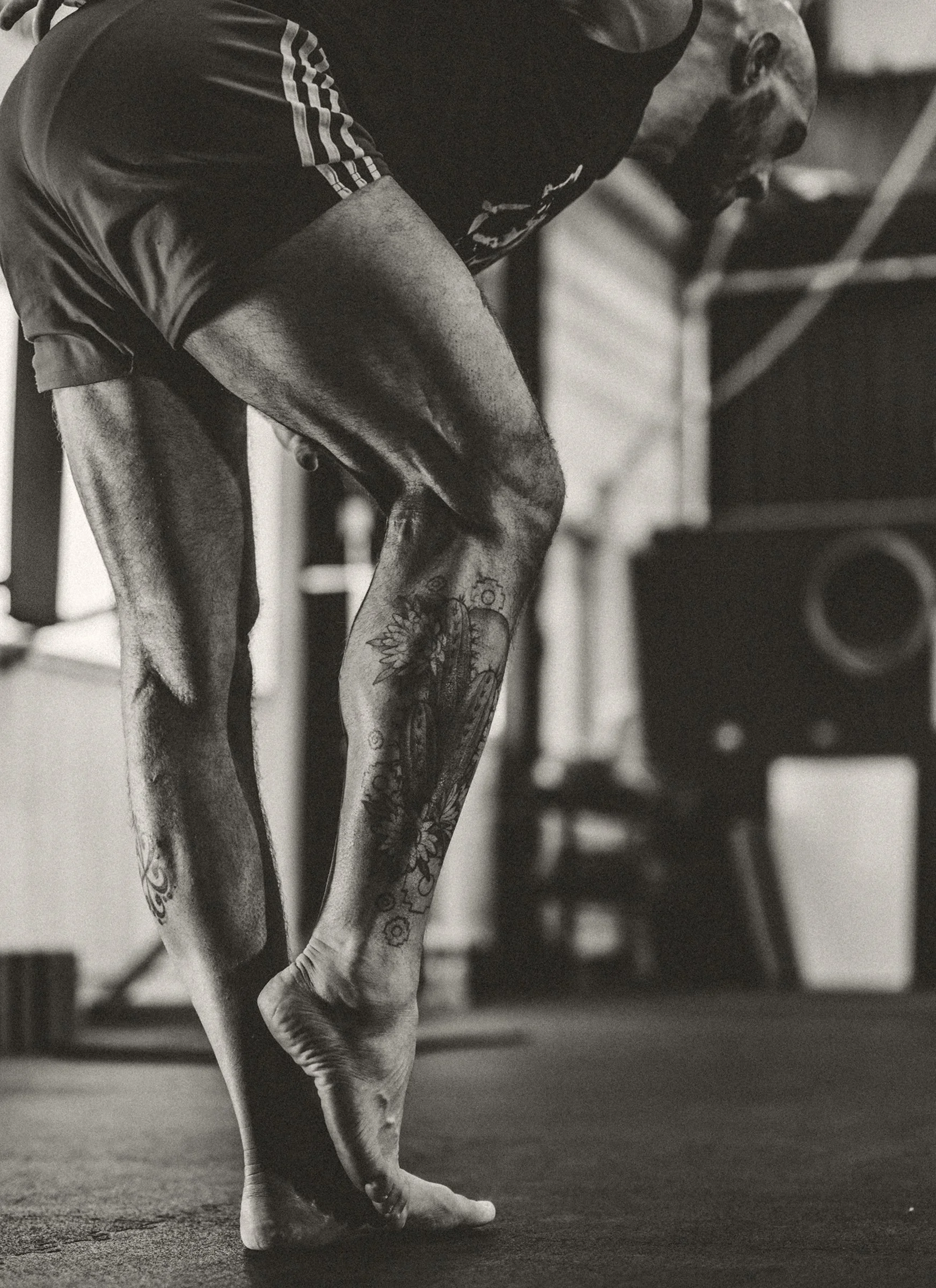 Close-up of a person standing on the tiptoes with bent knees in a gym, wearing shorts, showing tattoos on legs, with gym equipment in the background.
