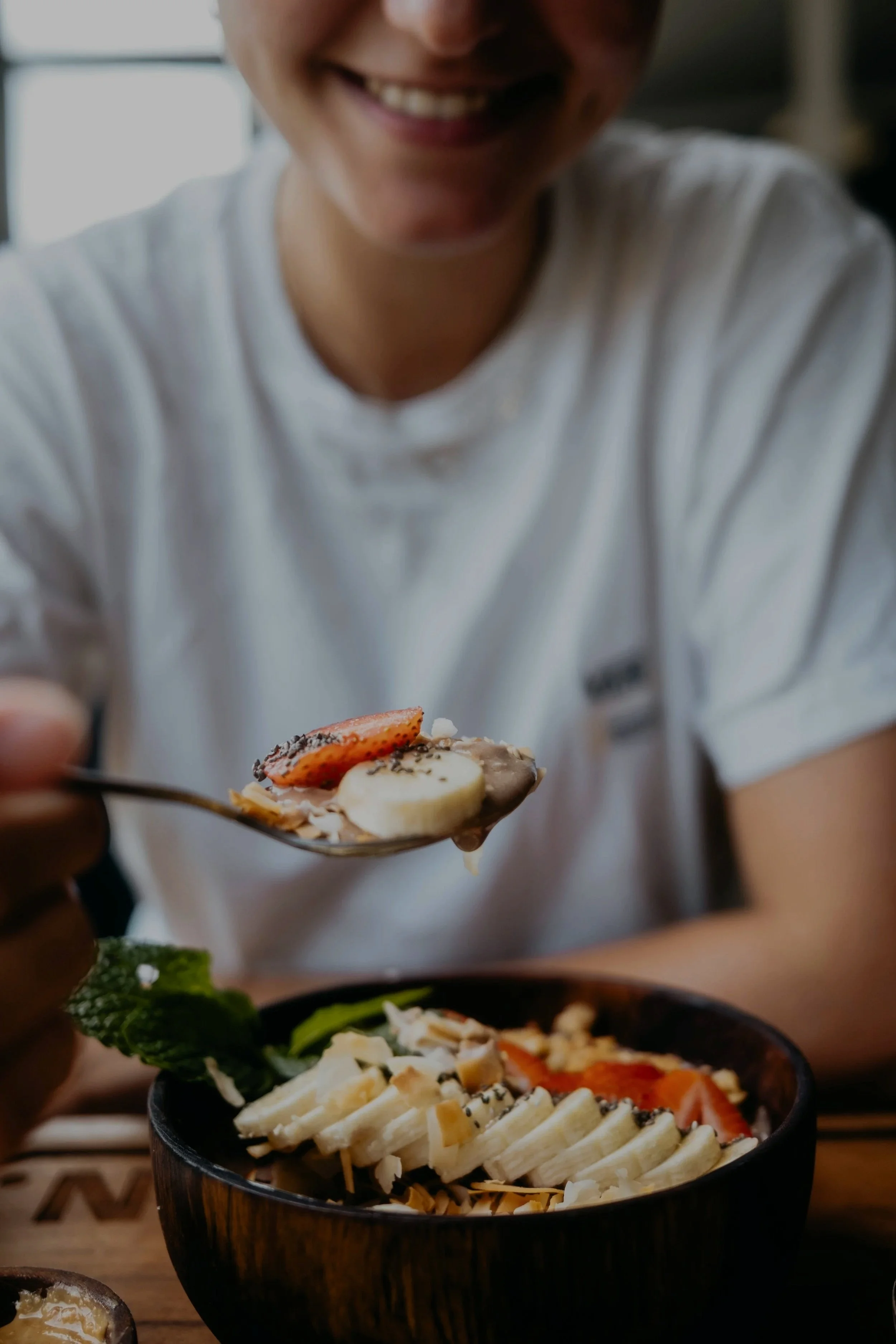 Person smiling and holding a spoon with a fruit-topped dessert over a bowl of sliced bananas, strawberries, and other fruits.