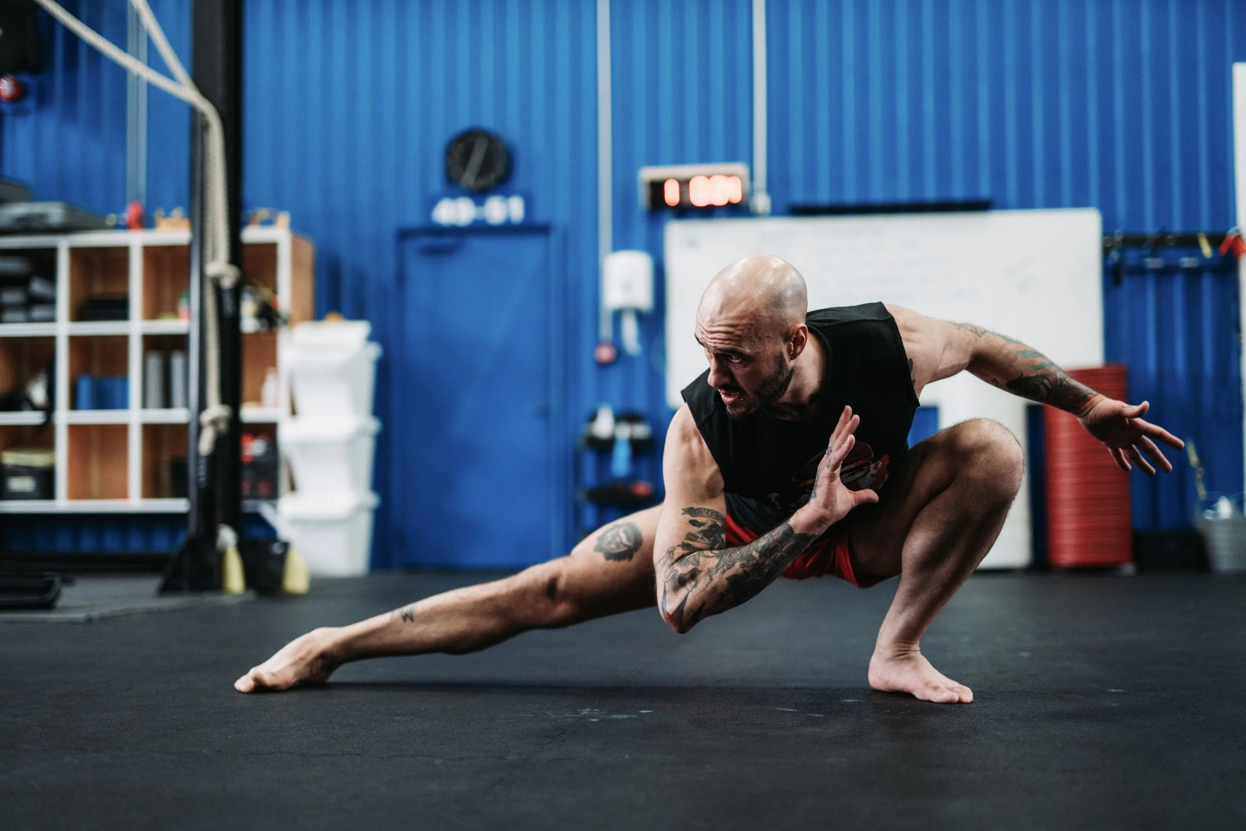 A man with tattoos practicing martial arts or fighting stance on a black gym floor in a gym with blue walls, equipment, and whiteboards.