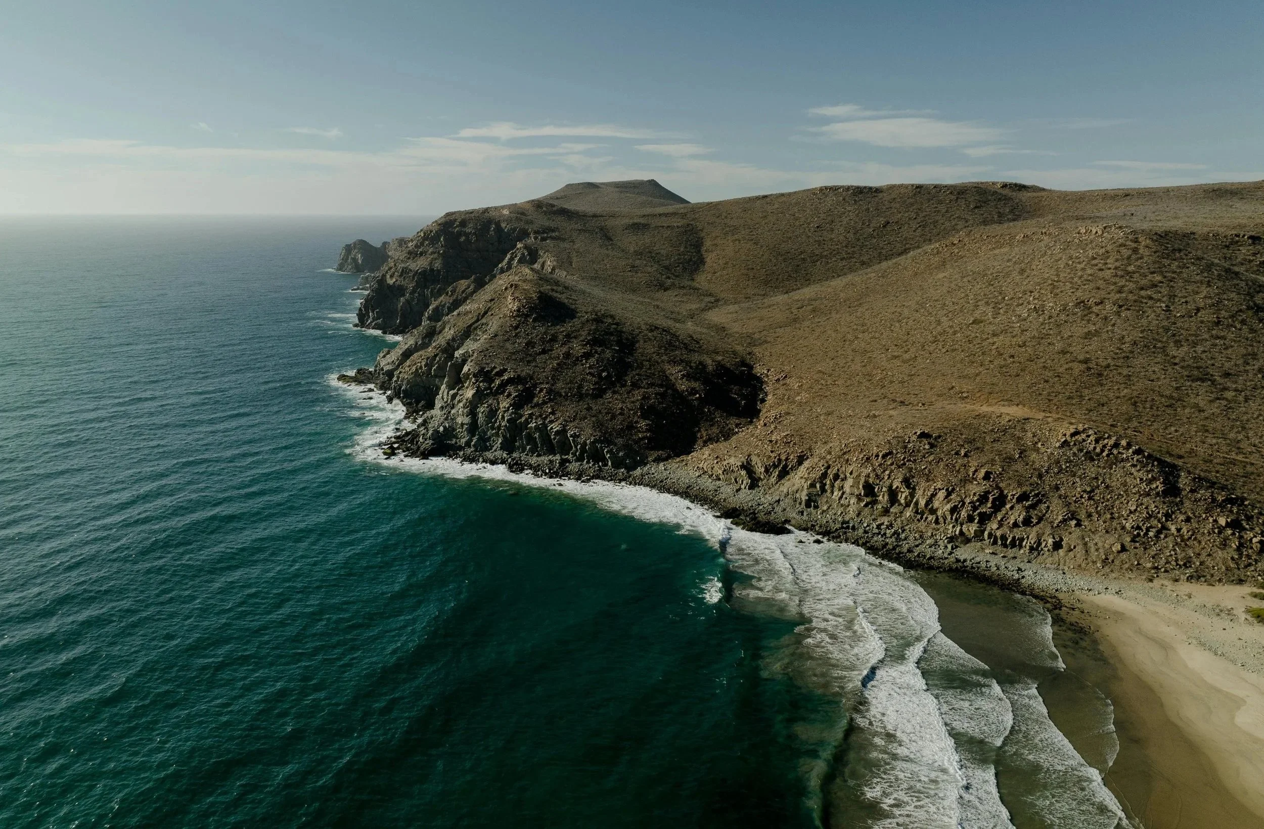 Aerial view of a rugged coastline with steep hills and cliffs meeting the ocean, with waves crashing onto a narrow beach.