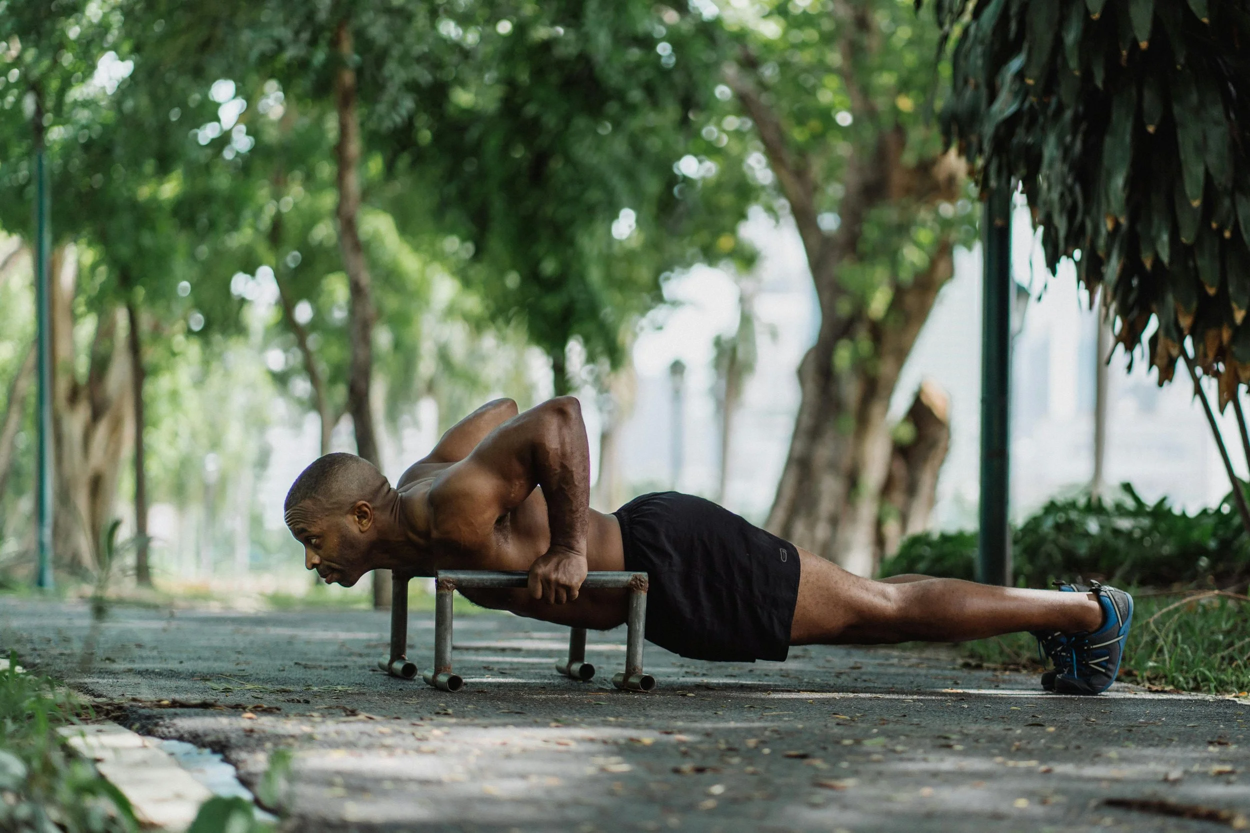 A shirtless man doing push-ups with his arms on a push-up bar in a park surrounded by trees.