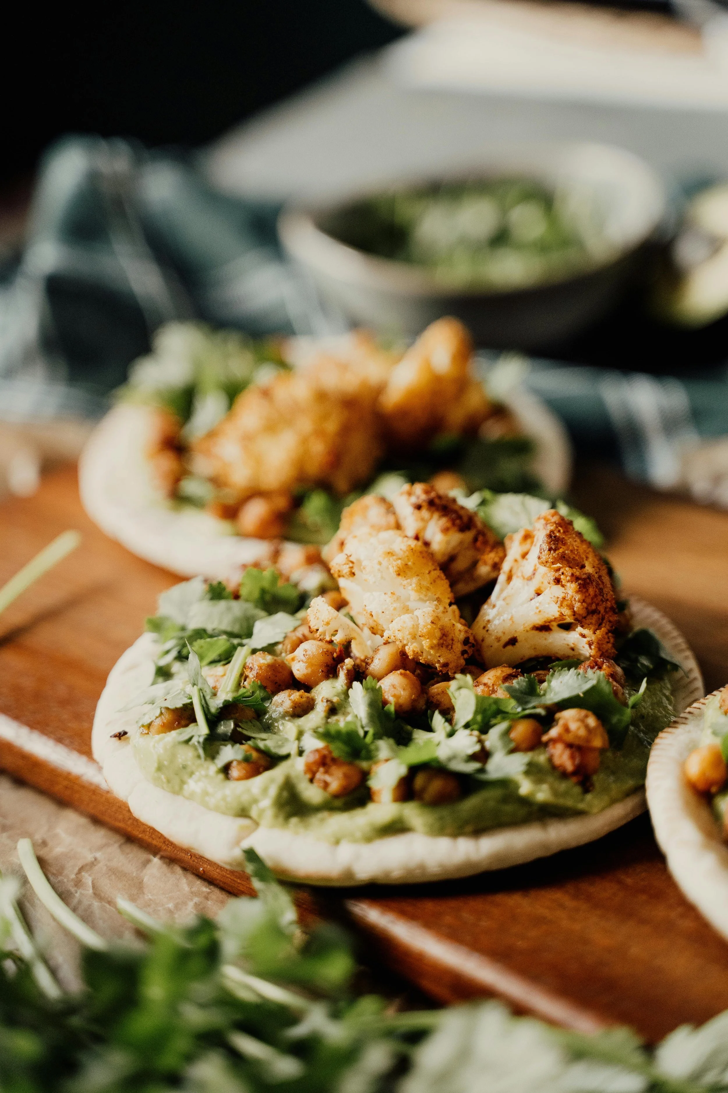 Close-up of flatbread topped with cauliflower, chickpeas, and green sauce, on a wooden surface.