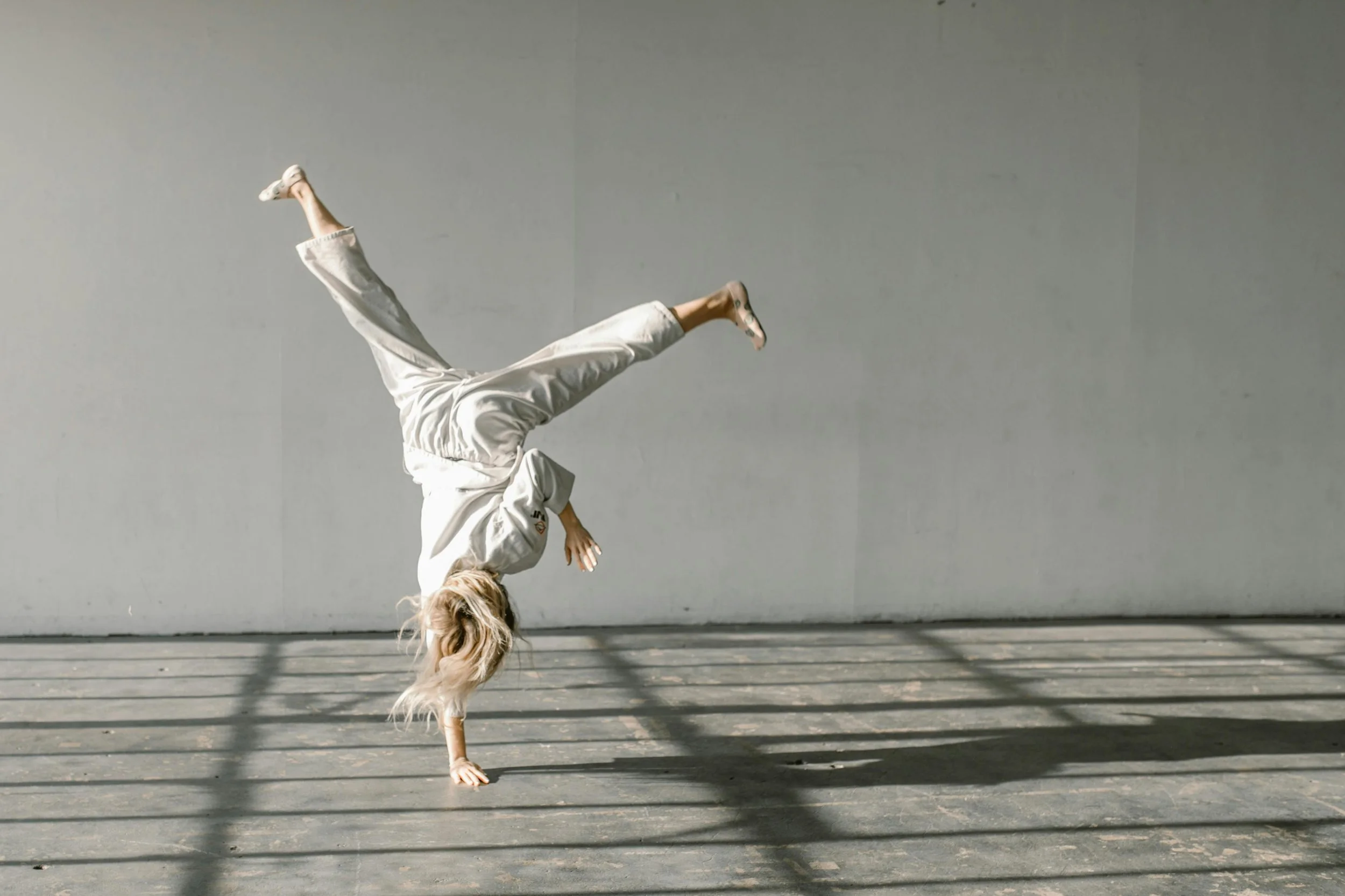 Person wearing a white outfit performing a handstand on gray floor with shadows and plain gray wall in background.