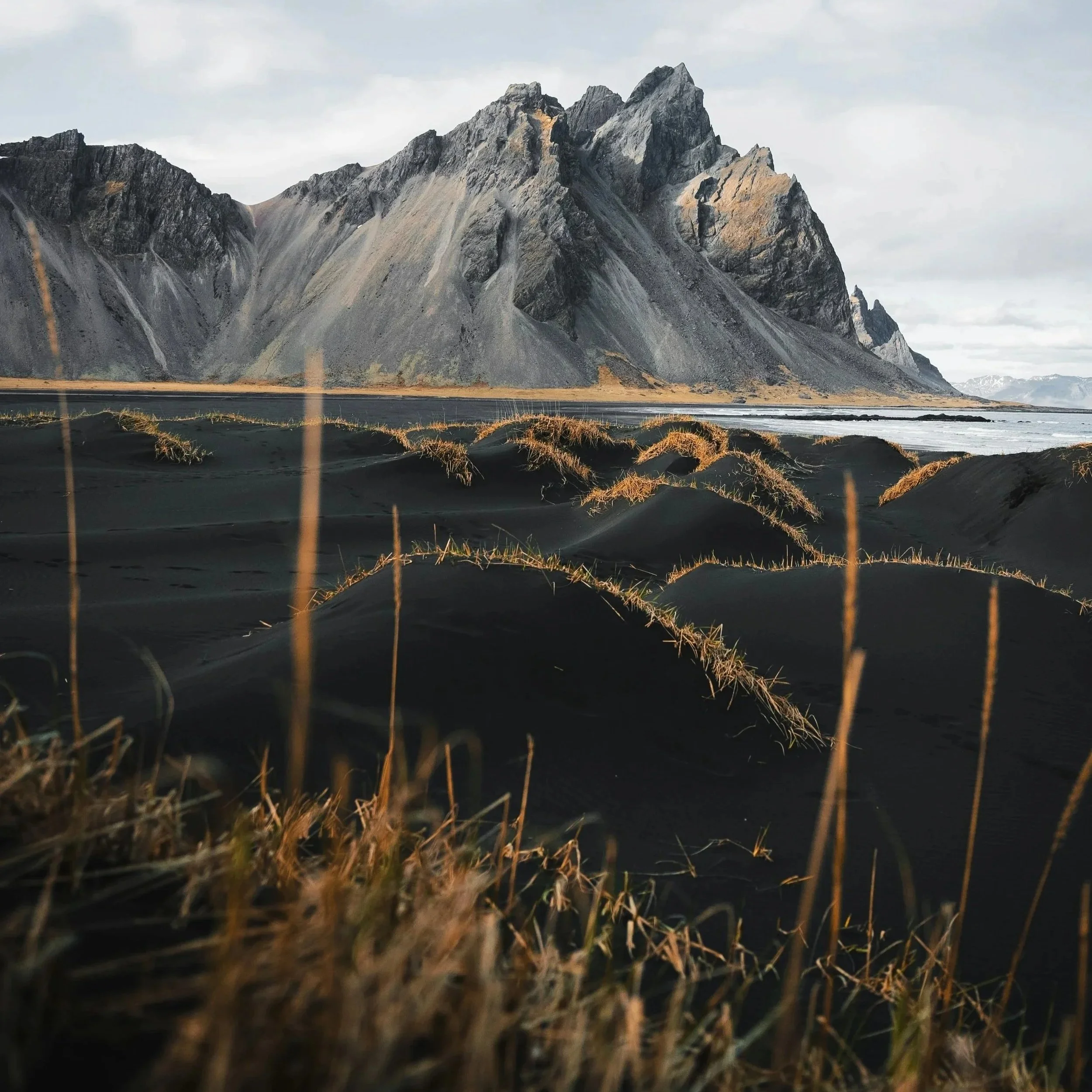Black sand dunes with sparse golden grass, rugged mountains in the background, and a cloudy sky.