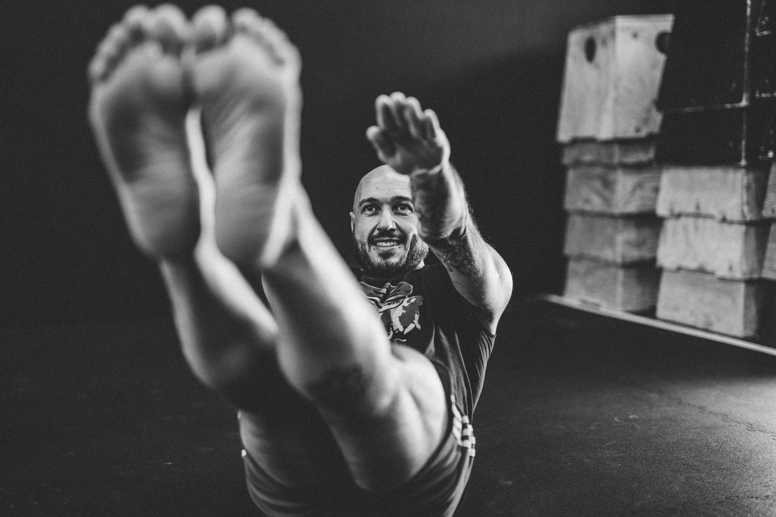 A man with a shaved head and beard exercises on the floor, laughing, with stacked wooden boxes in the background.