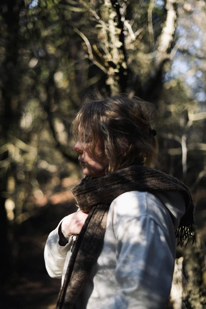A woman with curly hair wearing a brown scarf and a white jacket, standing outdoors among trees with sunlight filtering through the branches.