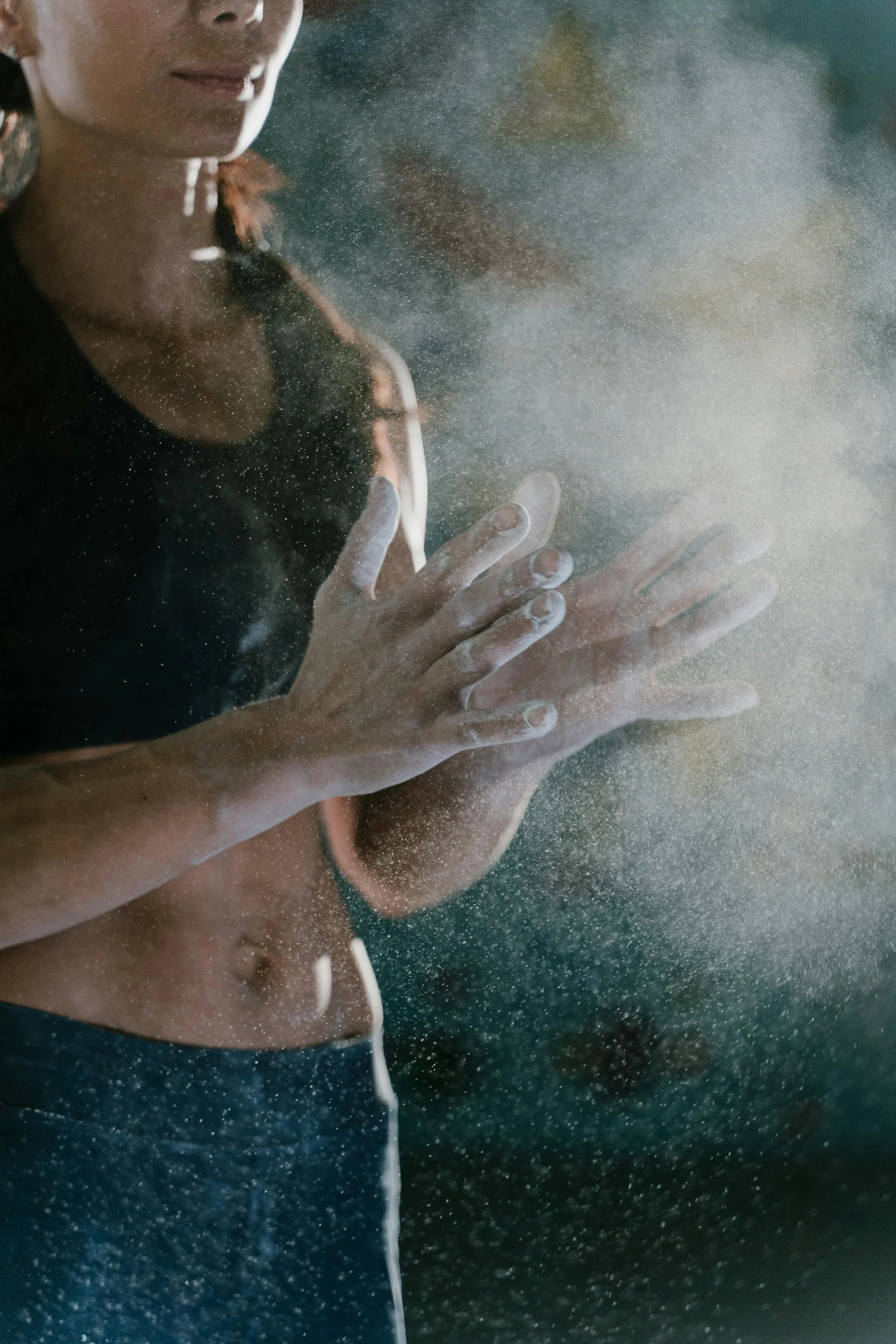 A woman with dark hair and wearing a black sleeveless top is clapping or rubbing her hands together in a cloud of dust or chalk powder, with a dark background.