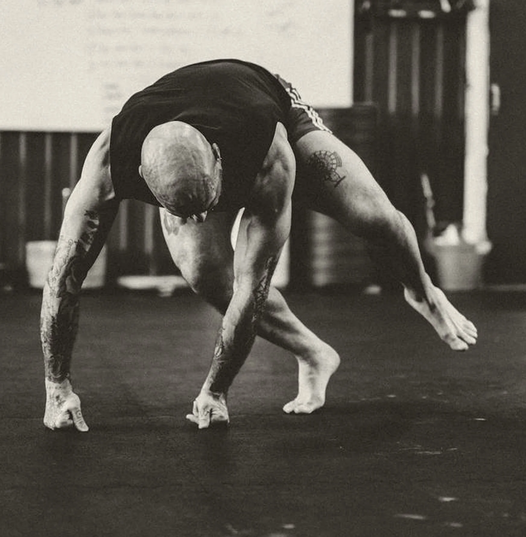 A tattooed man with a shaved head practicing a yoga pose, balancing on his hands and one foot, in a gym.