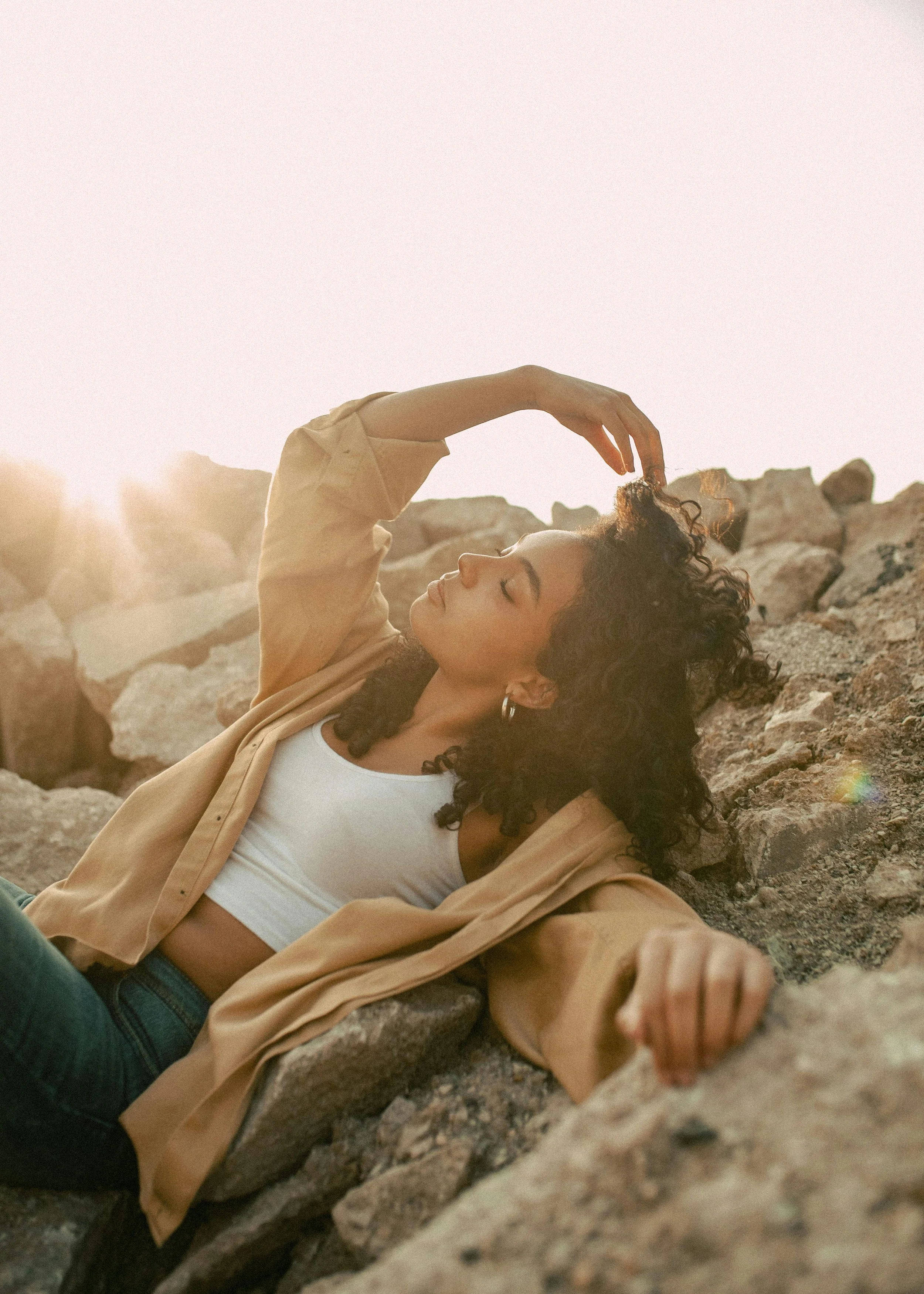 A young woman with curly dark hair, wearing a white crop top, beige jacket, and hoop earrings, is laying on rocks outdoors at sunset with her eyes closed and one arm raised above her head.