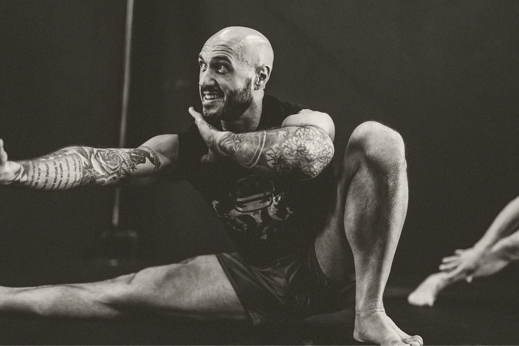 Black and white photo of a tattooed man with a shaved head doing a high kick in a martial arts or fitness class, smiling.