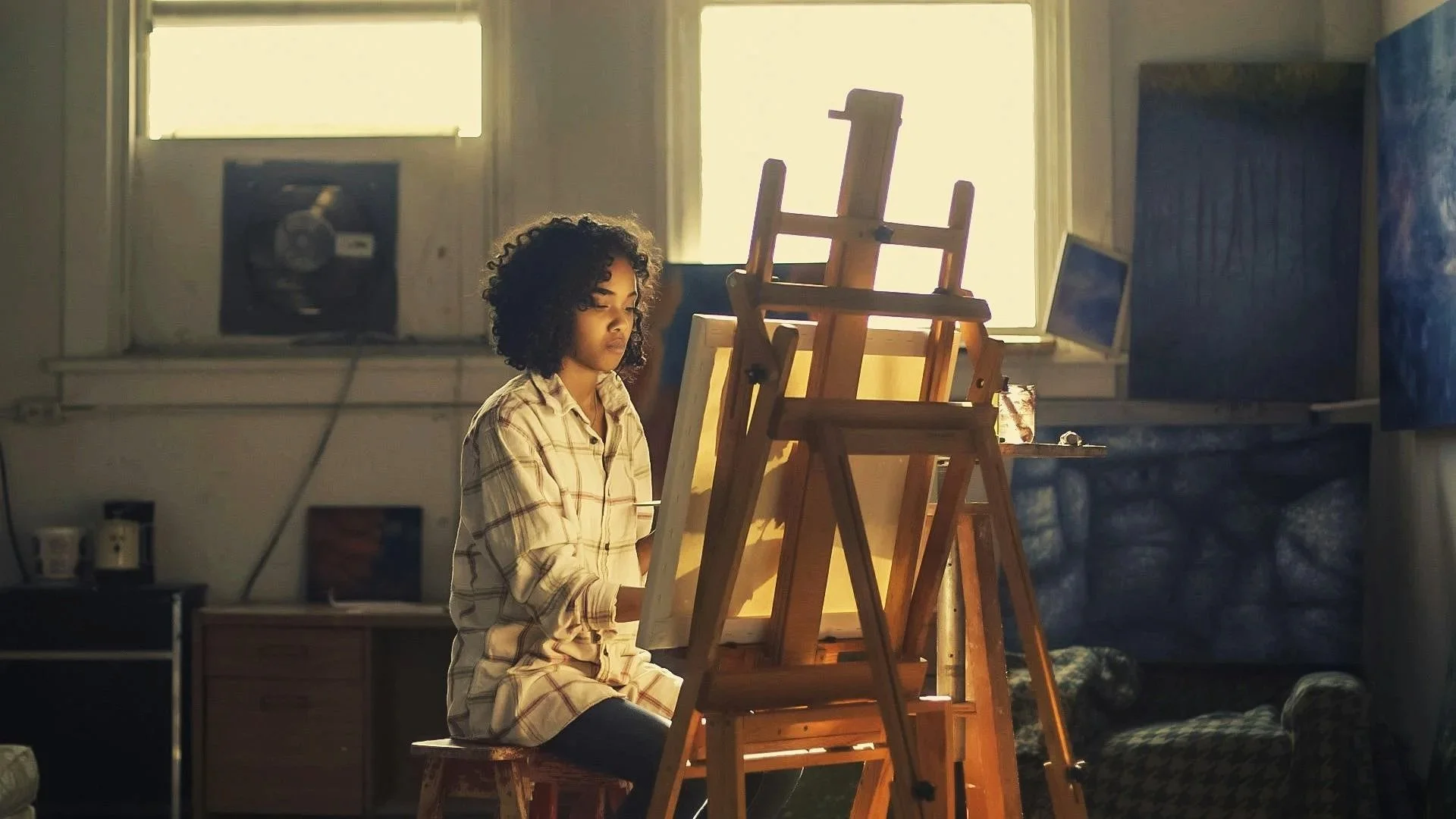 A young woman with curly hair sits on a stool in front of multiple wooden easels, painting or drawing in a well-lit room with large windows and various art supplies.