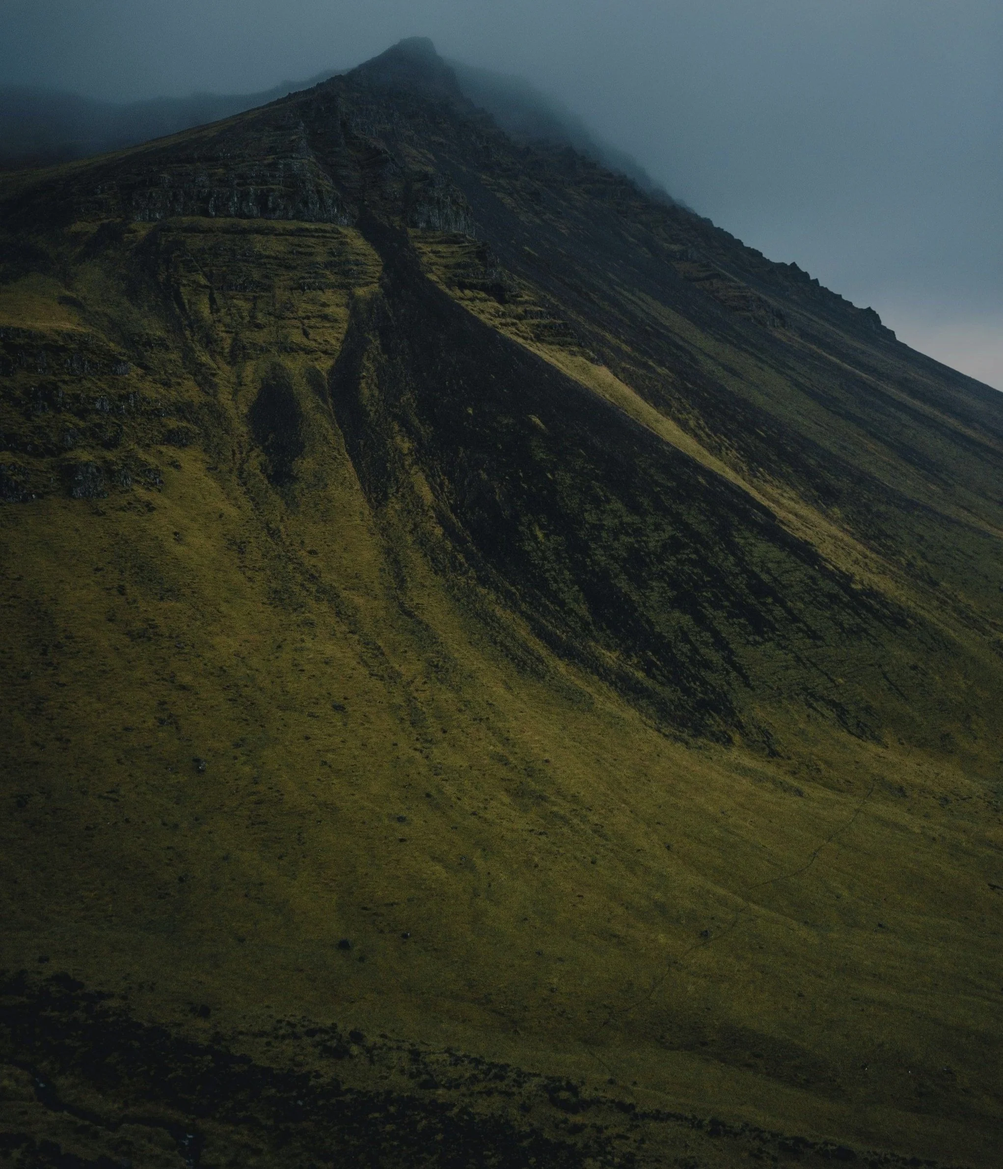 A foggy mountain with steep, grassy slopes and a rugged peak at the top.